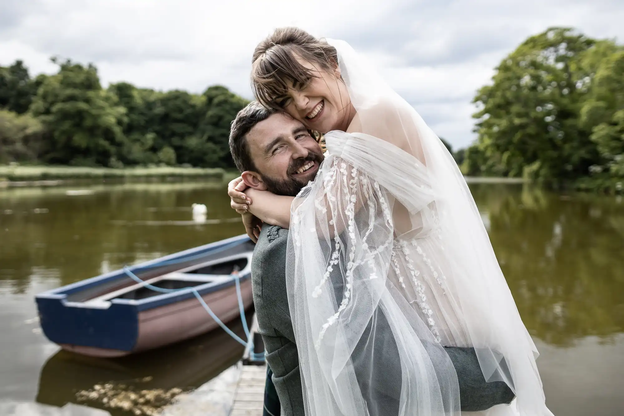 A smiling bride in a sheer, beaded veil and dress is joyfully lifted by a bearded groom in a grey suit on a jetty beside a rowing boat, with a calm lake and lush green trees in the background under a cloudy sky.