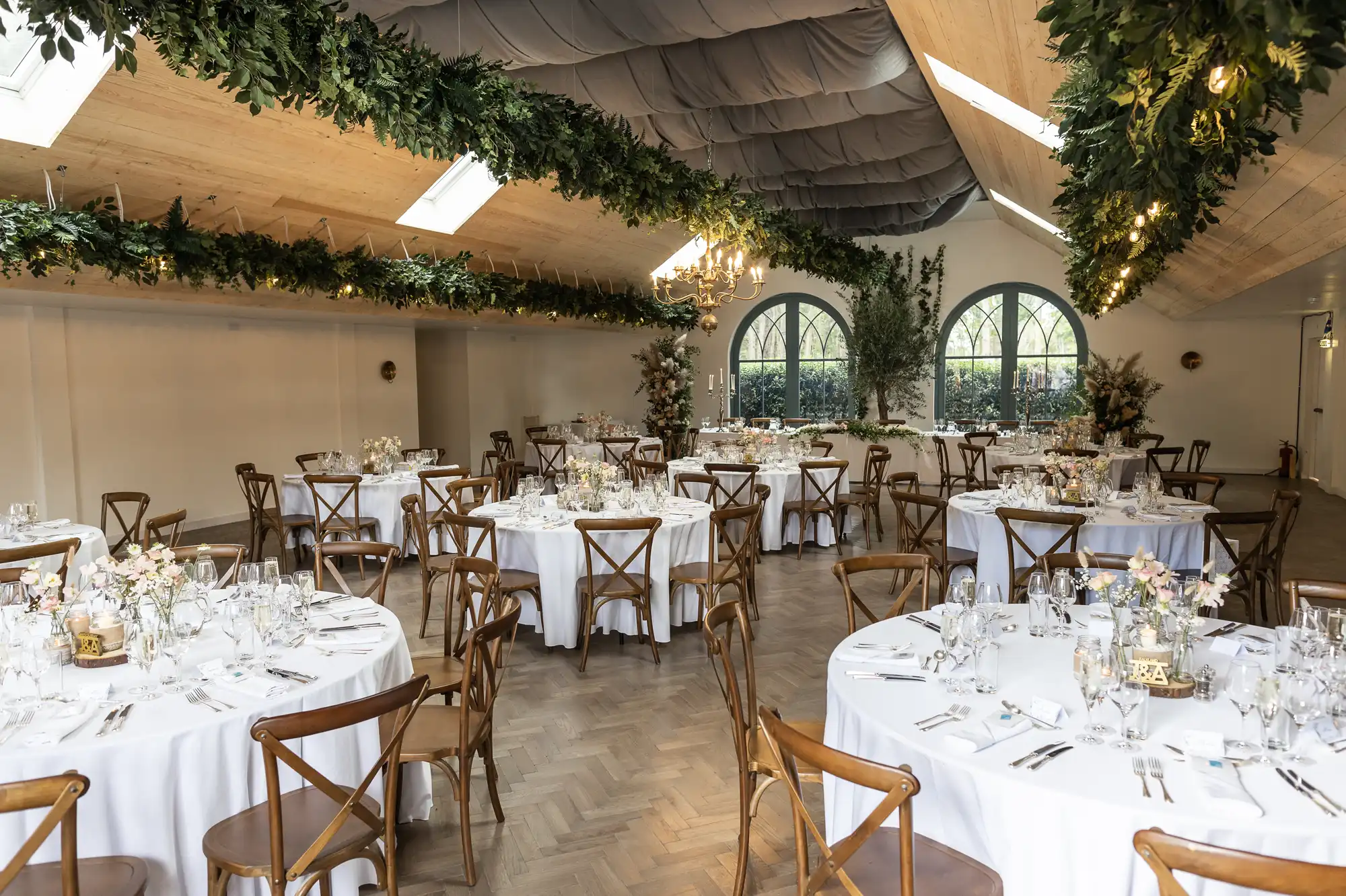 Elegant wedding reception hall with round tables covered in white cloths, set with glassware and floral centrepieces. Wooden chairs surround the tables. Greenery and fairy lights decorate the ceiling, and large arched windows let in natural light.
