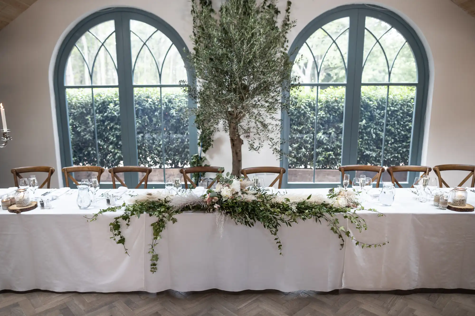 A long, white-clothed table is elegantly set for a formal event. Greenery and white flowers cascade over the centre. Wooden chairs line the table, with large arched windows and an indoor tree creating a bright, natural backdrop.