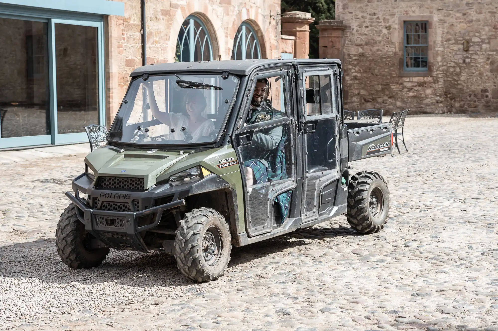 A green off-road utility vehicle drives on a cobblestone courtyard beside a stone building with arched windows. Two people sit inside the vehicle, one driving and one in the passenger seat. The vehicle has large tyres and an open cargo area at the back.
