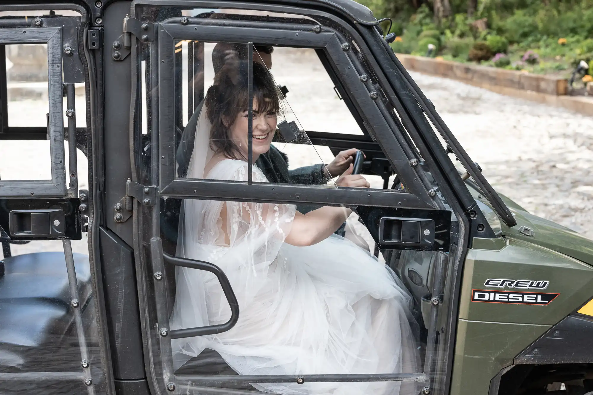 A bride in a white wedding dress and veil smiles whilst sitting in the passenger seat of a green utility vehicle labelled "DIESEL CREW." Another person, partially obscured, drives. The scene is outdoors with flowers and greenery in the background.