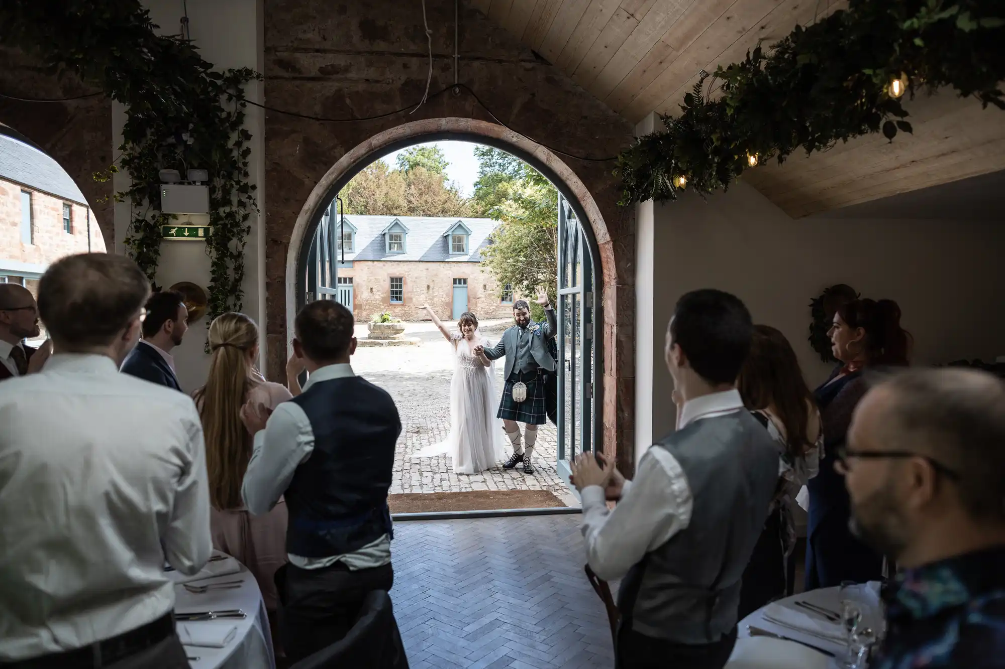 A bride and groom enter a rustic venue through a large arched doorway, greeted by standing guests who clap and smile. The indoor space has wooden ceilings and greenery, whilst outside, there’s a cobblestone courtyard and brick buildings.