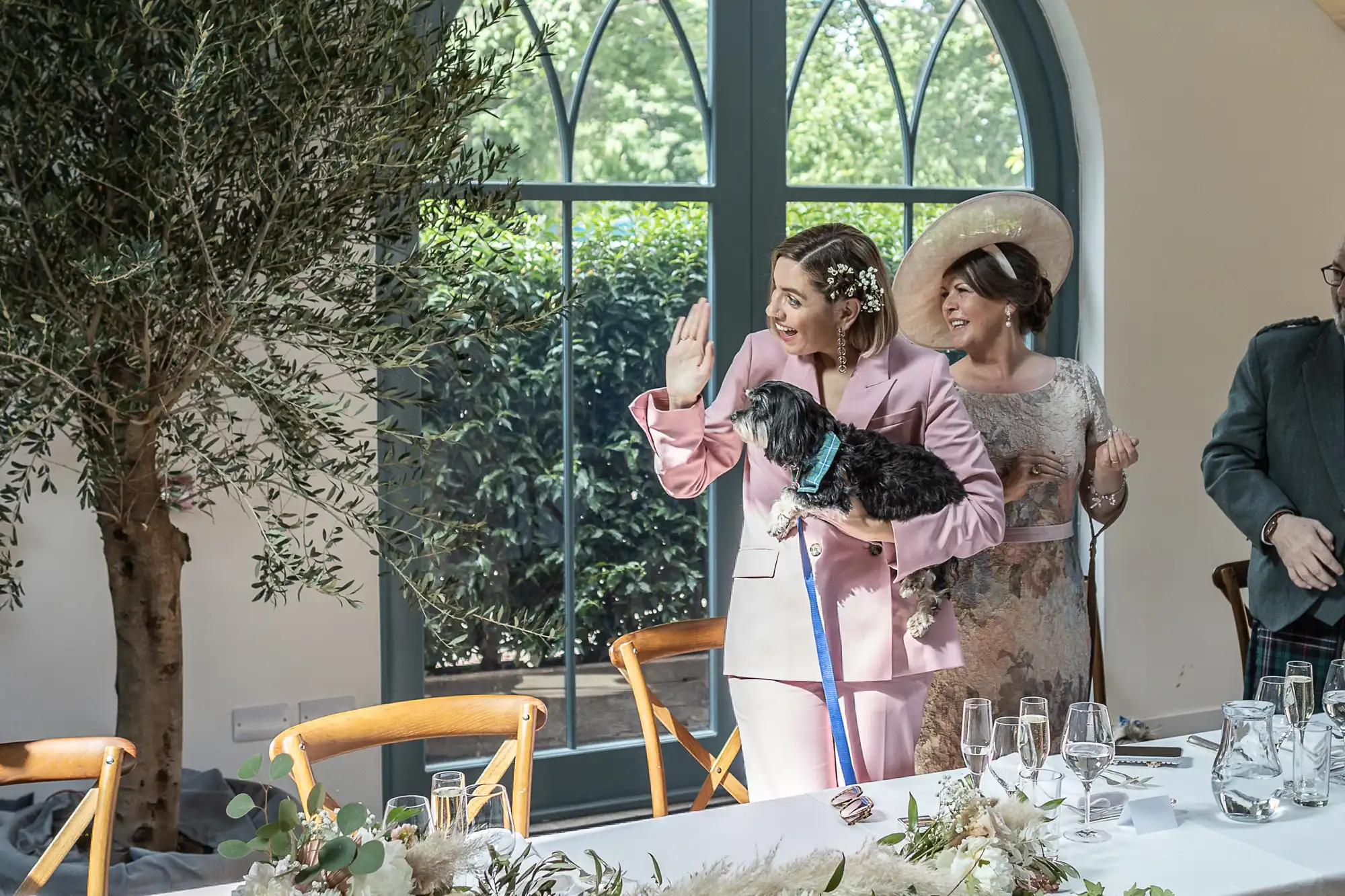 A smiling woman in a light pink suit holds a small black dog and waves. Beside her, an older woman in a floral dress and large hat smiles. They stand by a table set for a meal, near large arched windows and an indoor tree, with greenery outside.