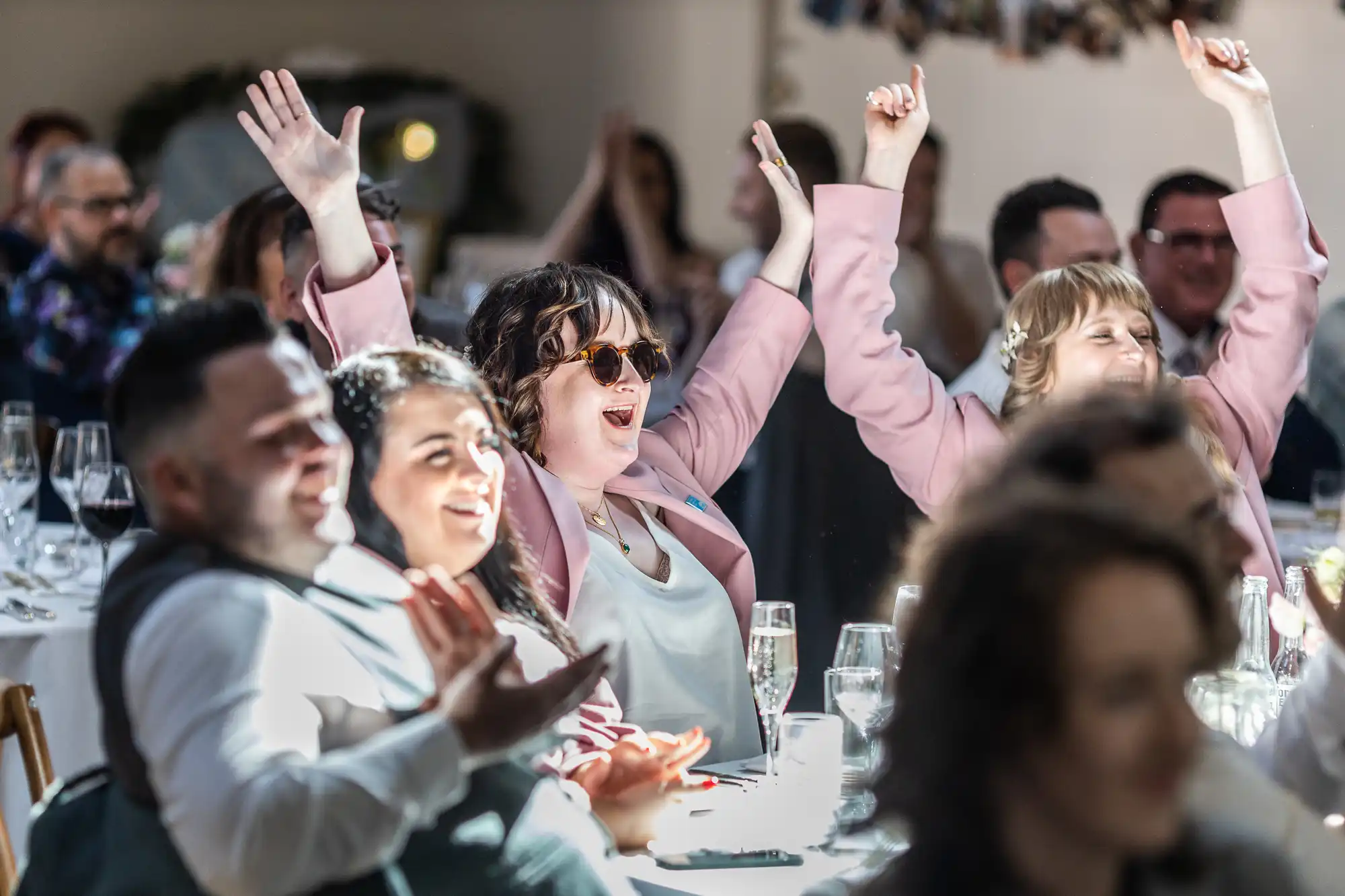 People seated at banquet tables smile and applaud. Two women in pink blazers, one wearing sunglasses, raise their arms joyfully. Sunlight brightens the lively atmosphere, with champagne glasses and blurred faces in the background.