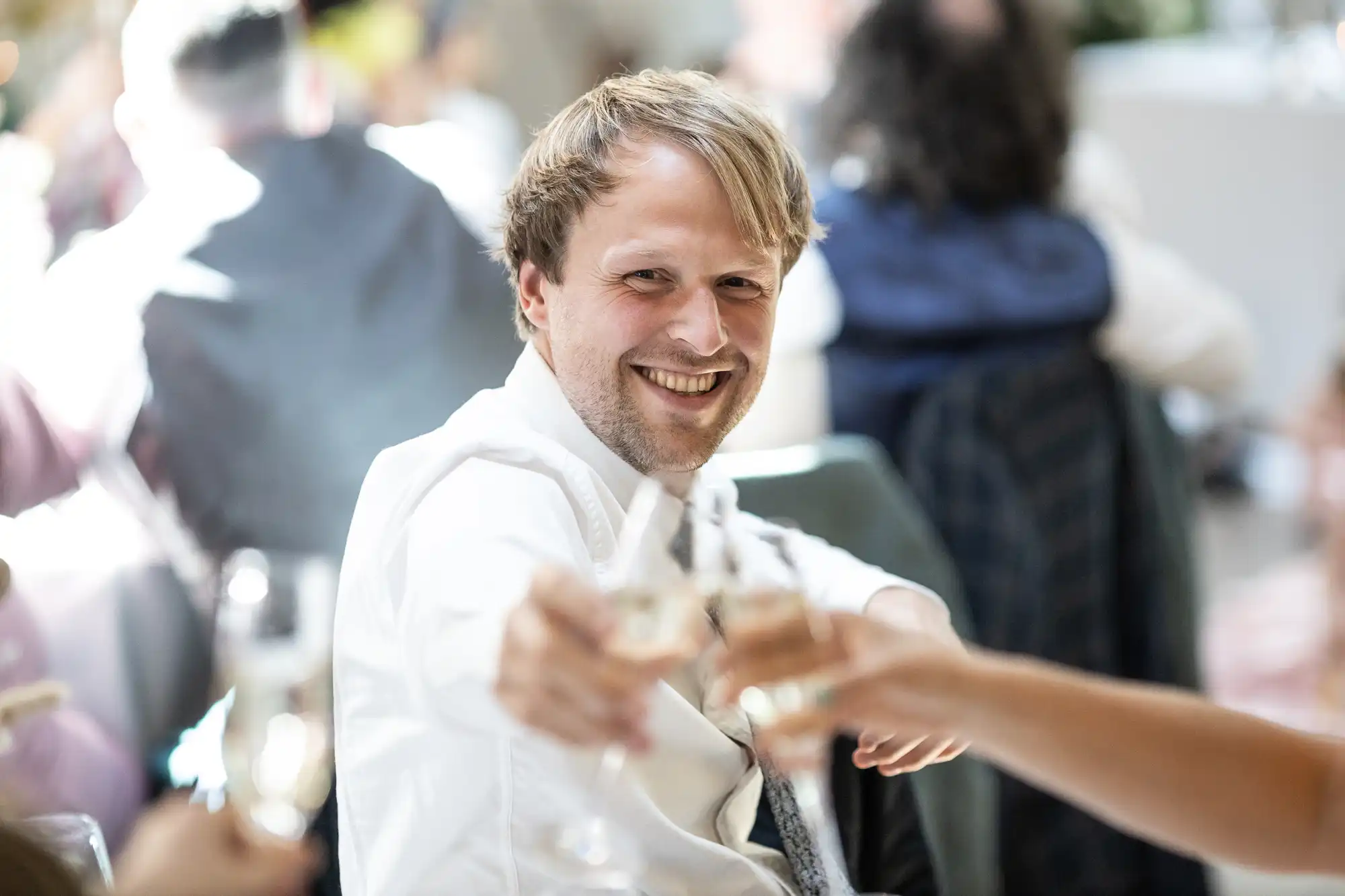 A man in a dress shirt and tie smiles warmly whilst toasting with others at a social event. He holds a glass, reaching forward to clink with outstretched hands. The background is softly blurred, showing more people and a bright, festive atmosphere.