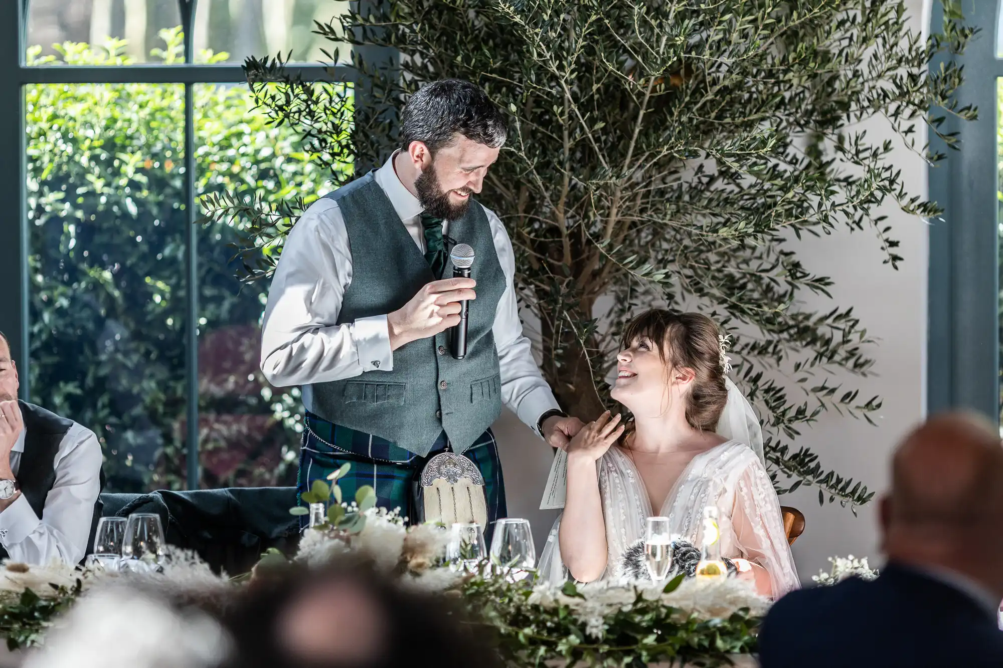 A man in a waistcoat and tie speaks into a microphone, smiling at a seated woman in a white dress who looks up at him, smiling. They are at a decorated table with flowers and glassware, with sunlight and greenery visible through large windows behind them.