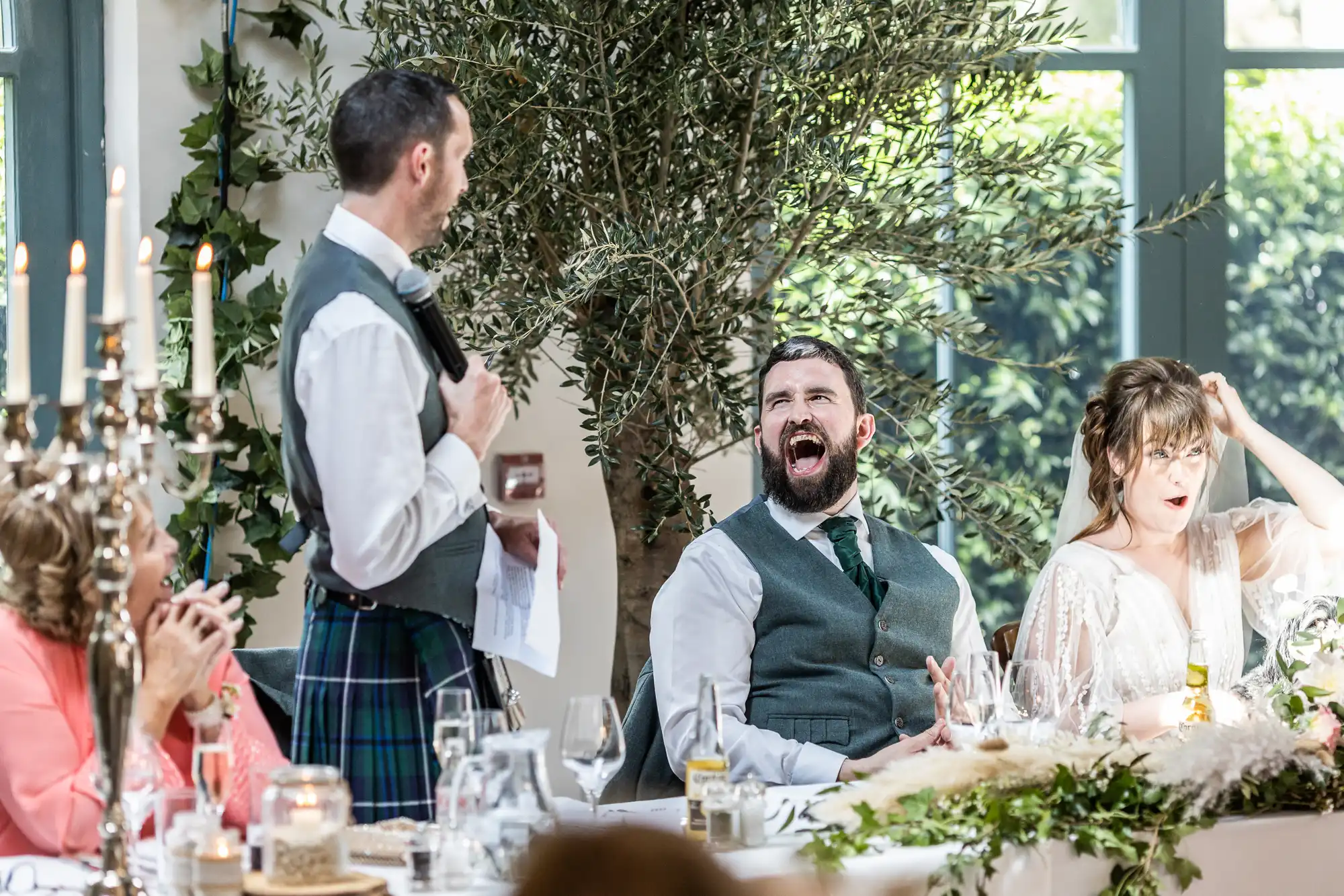A man in a kilt stands giving a speech with a microphone at a wedding table. Next to him, a bearded man laughs heartily, while a woman in a white dress wipes tears from her eyes. The table is decorated with greenery and candles.