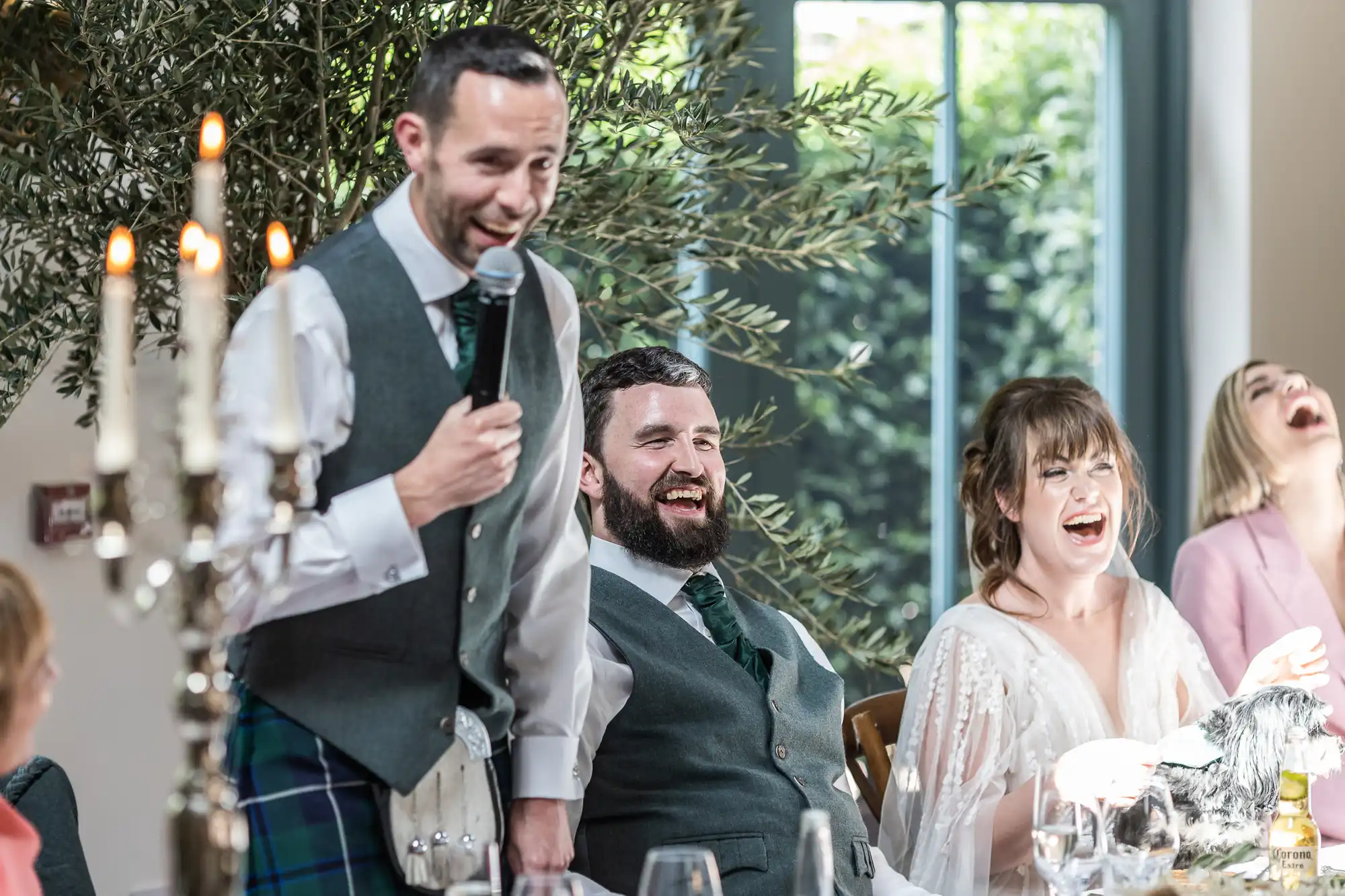 A man in a grey waistcoat and tartan holds a microphone, smiling as he gives a speech at a wedding table. Next to him, a groom, a bride in a white dress, and a woman in pink laugh joyfully. A candelabrum and green foliage are visible in the bright room.