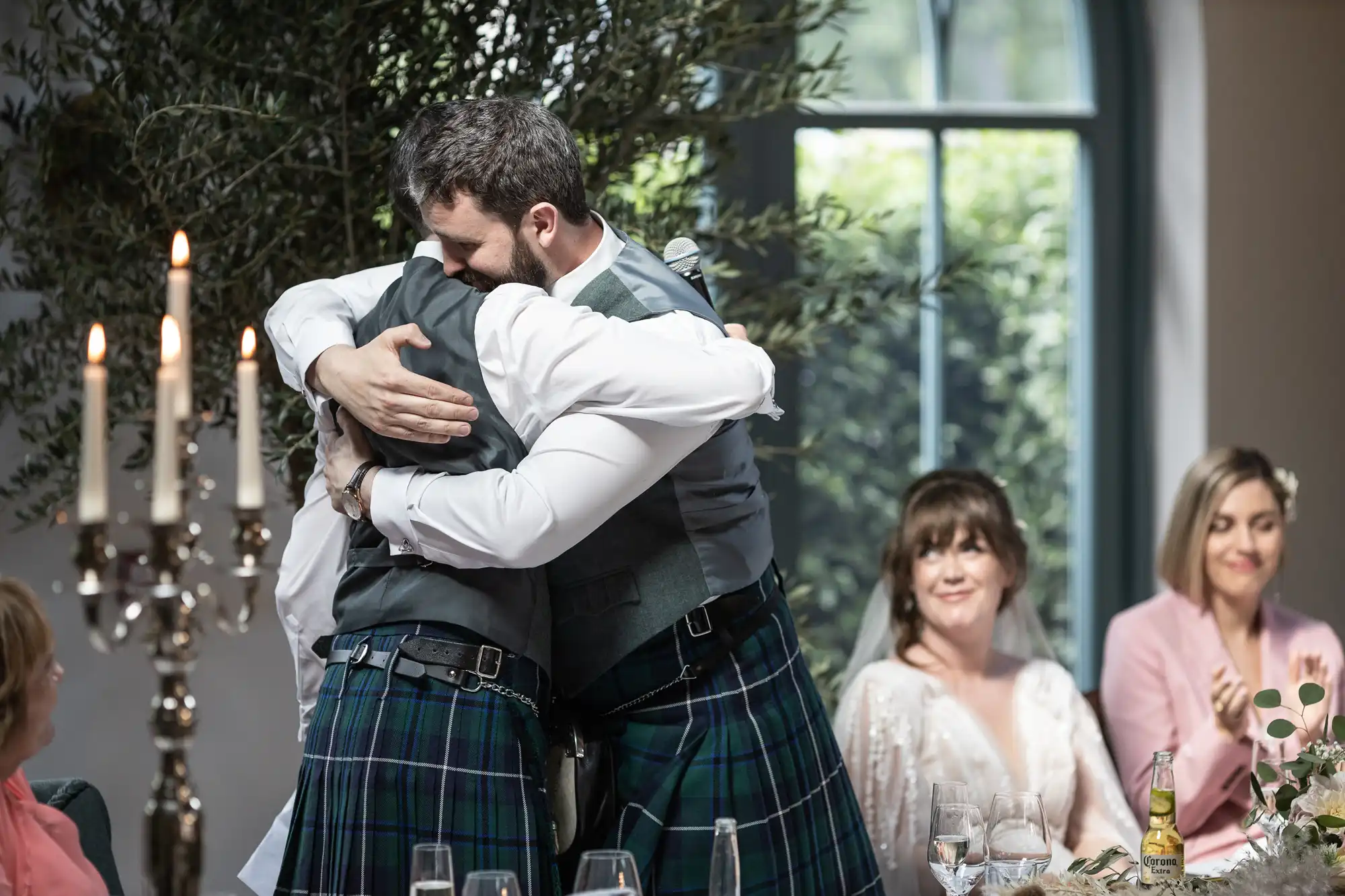 Two men in kilts hug warmly at a wedding reception, both wearing waistcoats and white shirts. Guests seated at a table, including smiling women in formal attire, look on. A candelabrum and greenery are visible, with sunlight streaming through a window in the background.