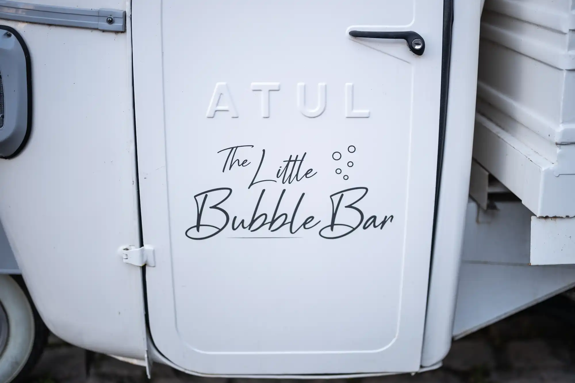 Close-up of a white vehicle door with embossed text "ATUL" and black script reading "The Little Bubble Bar" with three small bubbles illustrated between "Little" and "Bubble Bar." The door has a handle and black trim.