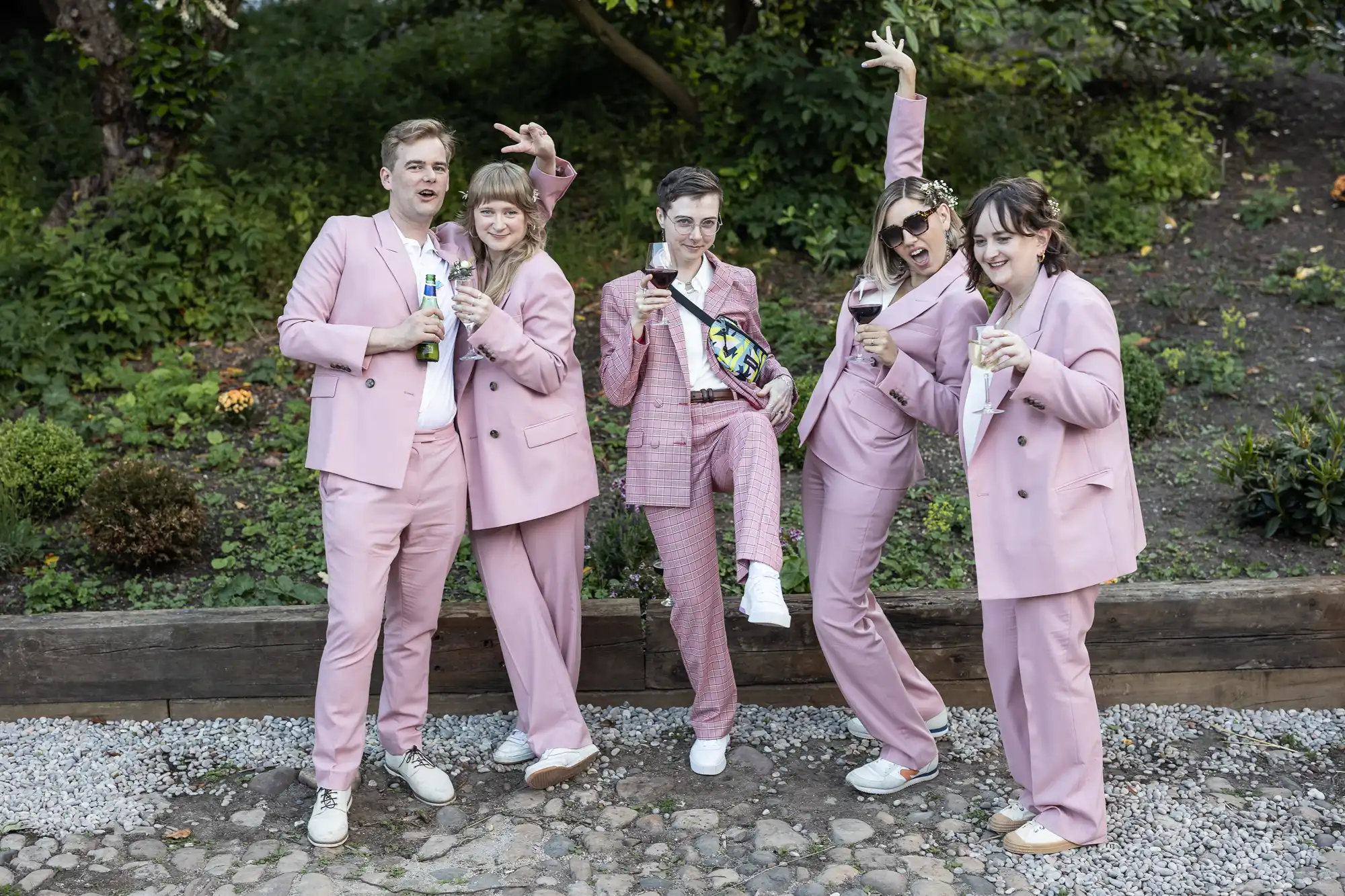 Five people pose outdoors in matching pale pink suits, white tops, and trainers. They hold drinks, smile, and make playful gestures, standing on a cobbled path with greenery and shrubs behind them, looking happy and celebratory.