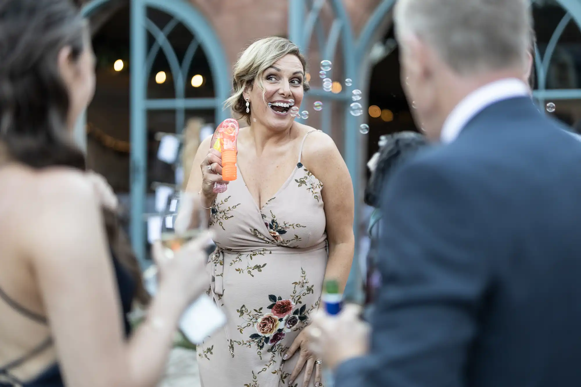 A smiling woman in a floral dress holds a bubble wand, blowing bubbles towards two people at an outdoor event. The background features arched windows and fairy lights, creating a festive atmosphere.