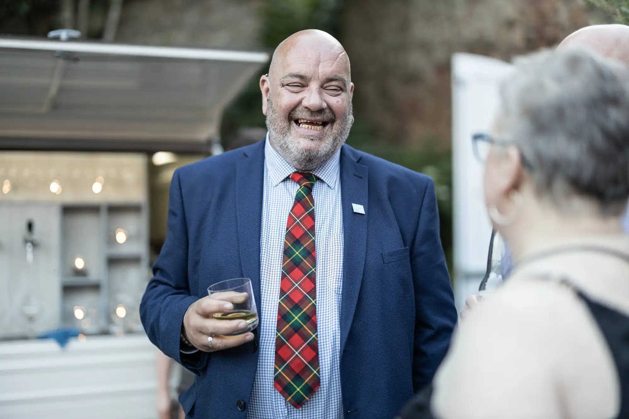 A bald man in a blue suit and red checked tie smiles whilst holding a drink at an outdoor event. He faces a woman with short grey hair and glasses. The background shows a white mobile bar with lights and blurred greenery.