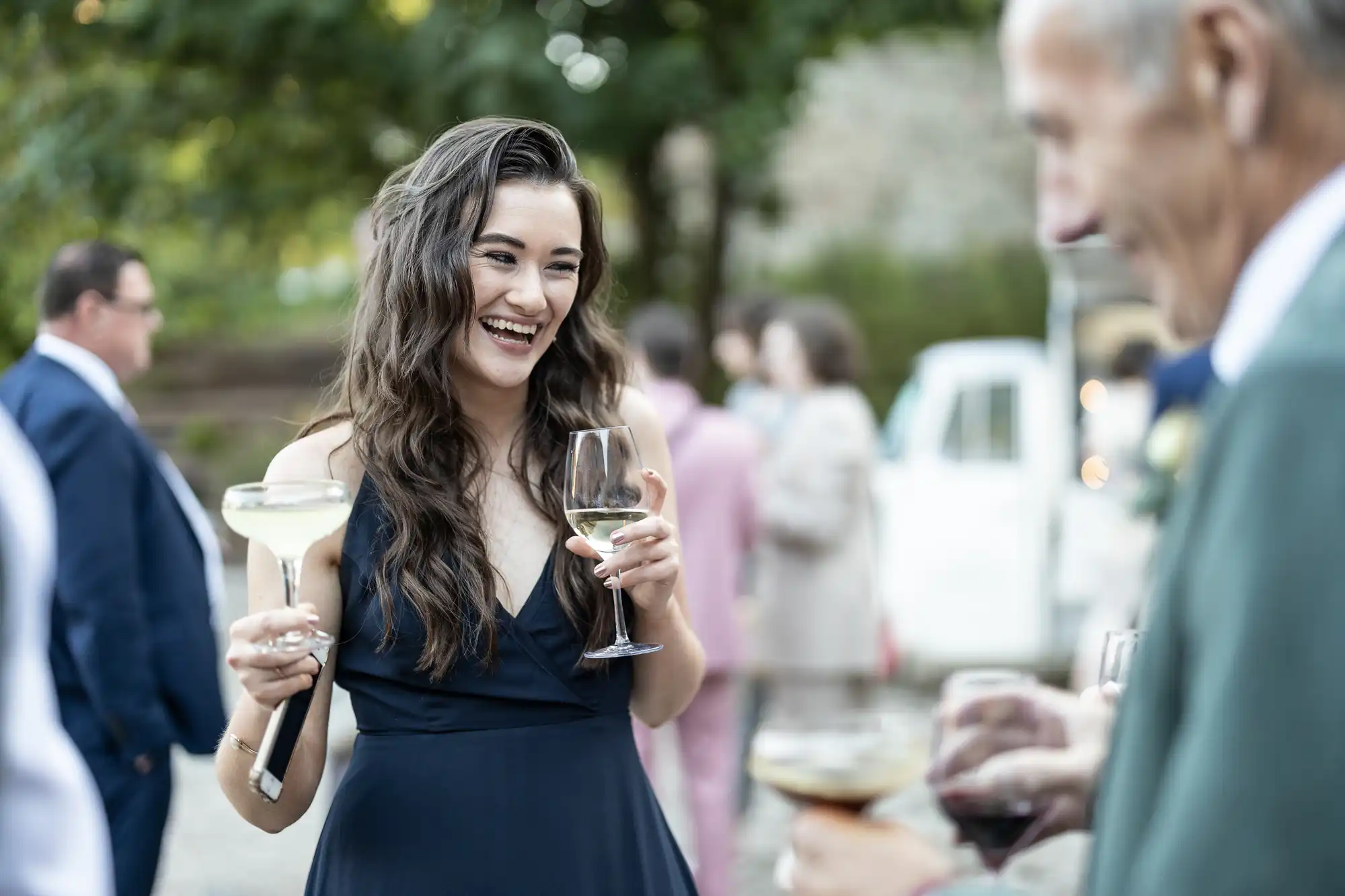 A smiling woman in a navy blue dress holds two wine glasses, one with white wine and another with a clear drink, whilst talking with an older man at an outdoor social gathering. Other guests and trees are blurred in the background.