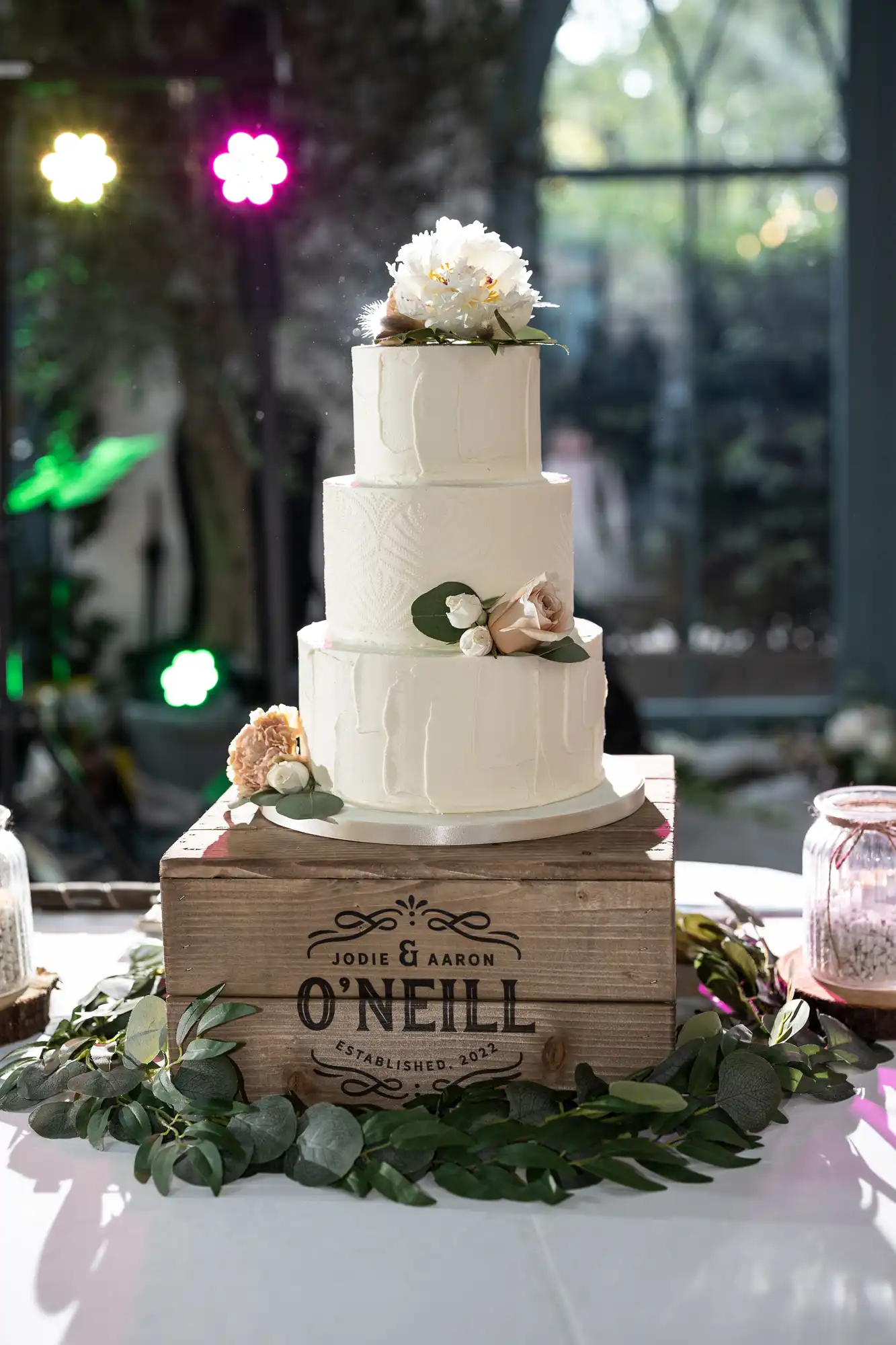 A white, three-tiered wedding cake decorated with white flowers and greenery sits on a rustic wooden crate that reads “Jodie & Aaron O’Neill Established 2023,” surrounded by eucalyptus leaves and small glass jars under soft lighting.