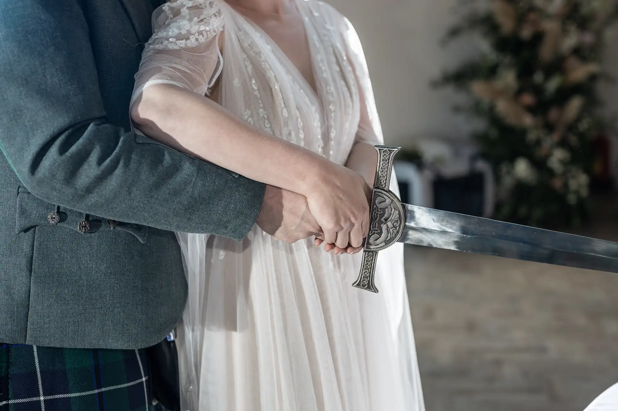 A couple in formal attire—one in a light, sheer dress, the other in a green jacket and checked kilt—stand close together, holding a large decorative sword with both hands. The background is softly blurred with greenery visible.