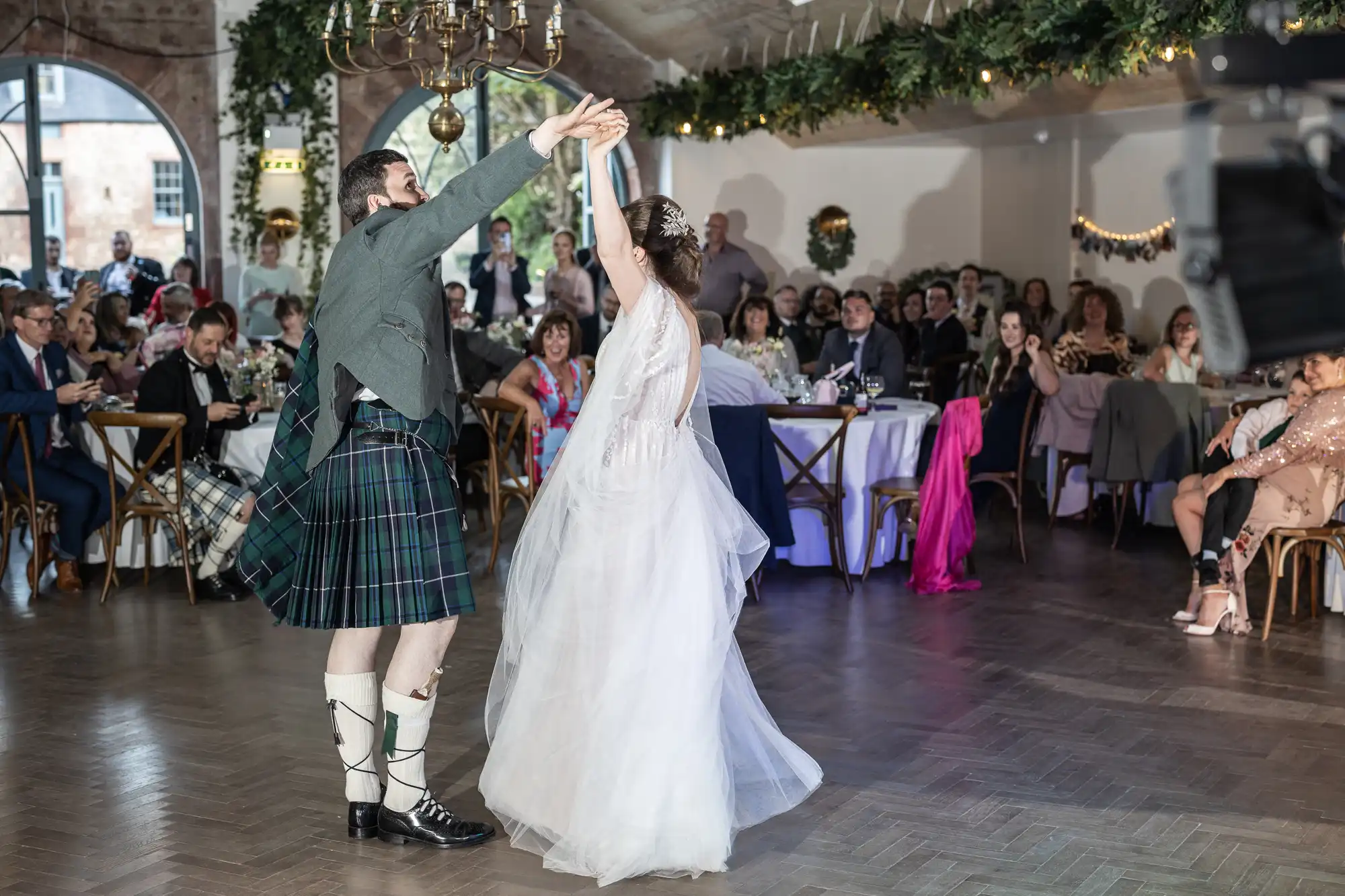 A bride in a white dress and a groom in a kilt dance together in a decorated hall, surrounded by seated guests watching and taking photos. The room is adorned with greenery and lights, creating a festive, joyful wedding atmosphere.