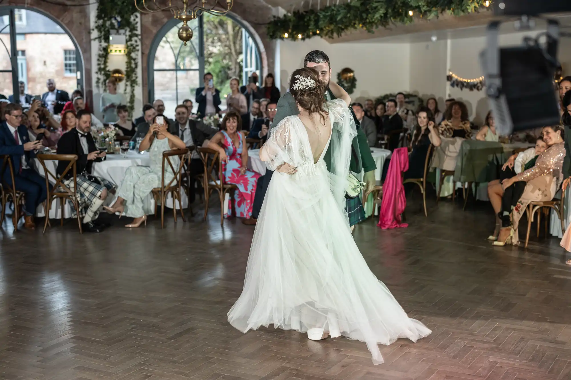 A bride and groom share their first dance in a decorated indoor venue, surrounded by seated guests watching and taking photos. The bride wears a flowing white gown, whilst the groom is in a dark suit. Greenery and chandeliers hang from the ceiling.