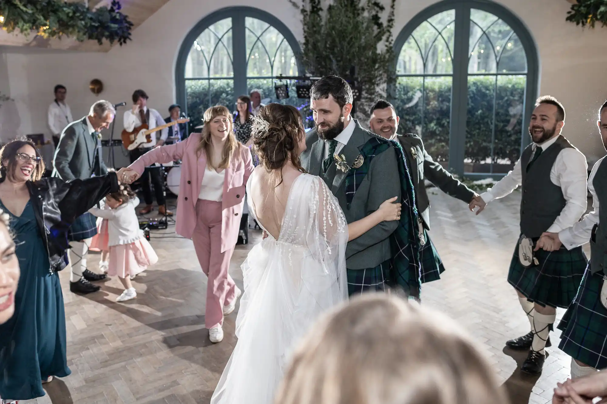 A bride in a white dress and a groom in a kilt dance together, surrounded by smiling guests in colourful attire holding hands in a circle, with a live band playing in the background and sunlight streaming through large arched windows.