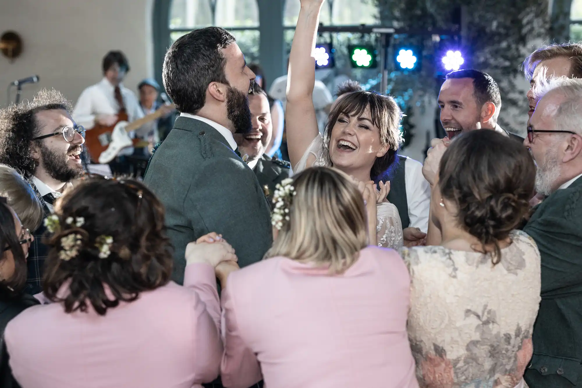A joyful bride and groom are surrounded by smiling guests in a lively group hug on the dance floor at a wedding reception. The bride raises her hand excitedly whilst people laugh, with a band playing in the background and colourful lights illuminating the scene.