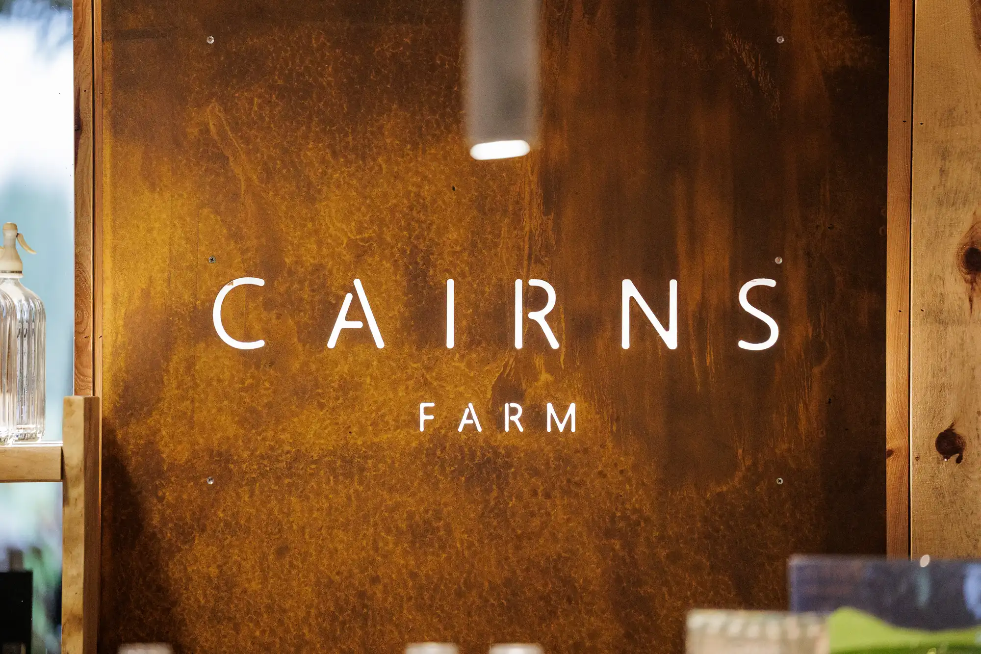 A close-up of a rustic, brown metal wall with the illuminated words "CAIRNS FARM" in modern white letters. A glass bottle is on a shelf at the left, and part of a wooden wall frames the image.