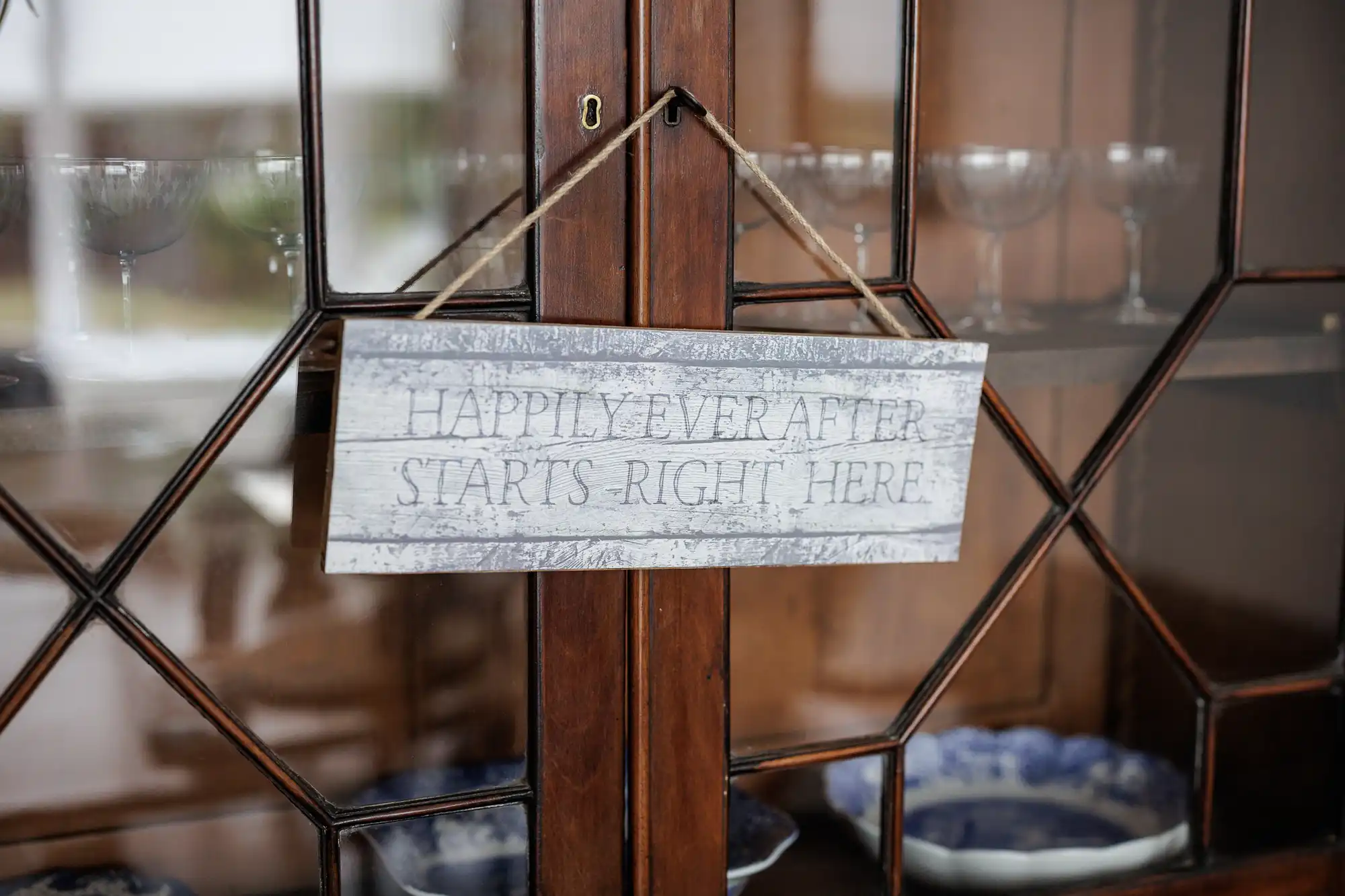 A rustic wooden sign reading "Happily Ever After Starts Right Here" hangs by twine on a glass-fronted wooden cupboard, with blue and white crockery and glassware displayed inside.
