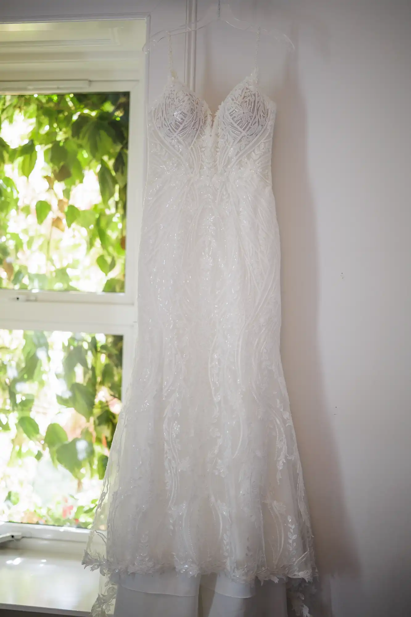 A white lace wedding dress with intricate patterns and a fitted bodice hangs on a hanger against a white wall. Sunlight filters through a nearby window with green leaves outside, softly illuminating the delicate fabric.
