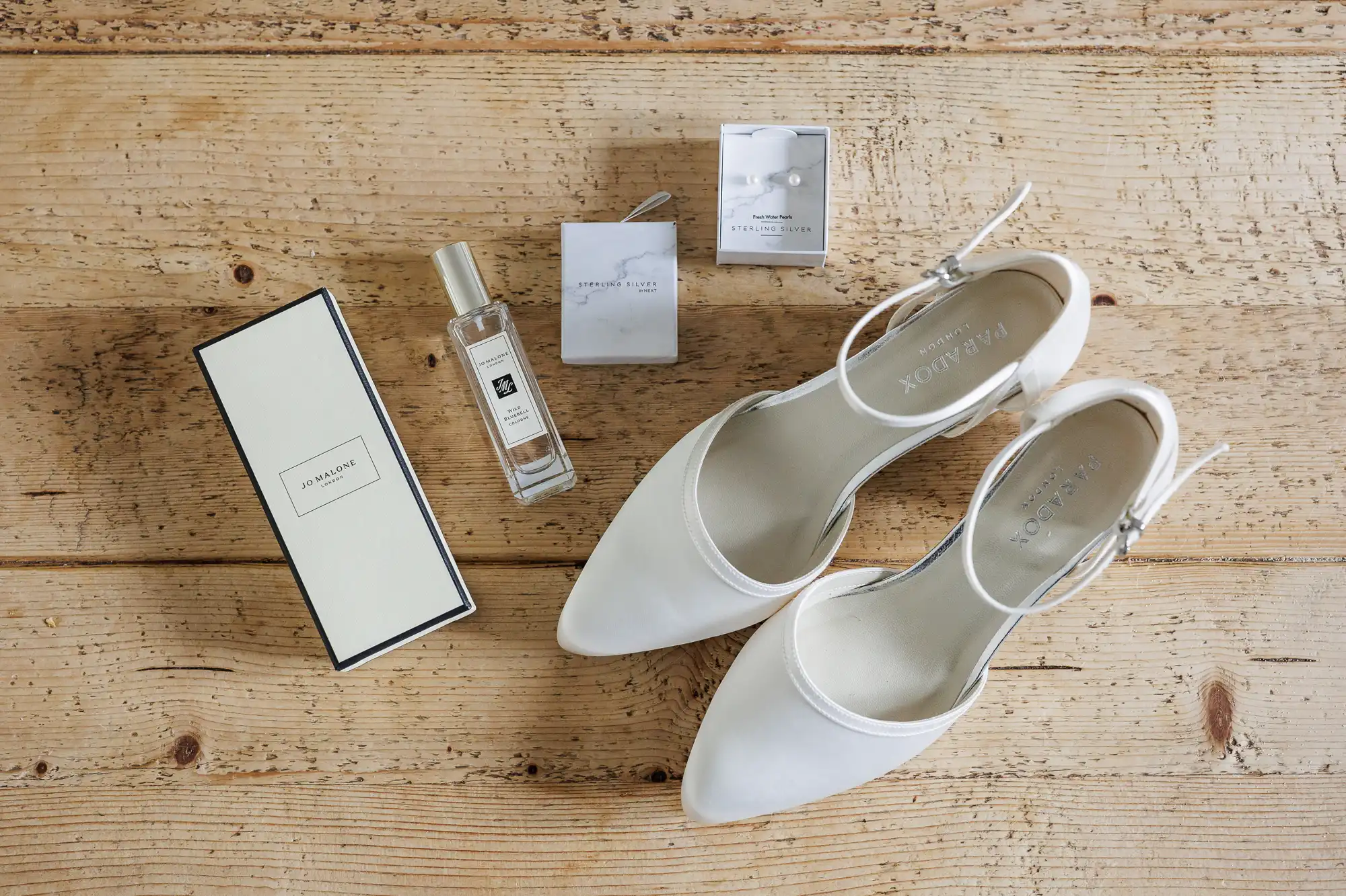 A pair of elegant white heels, a rectangular perfume bottle with a cream-coloured box, and two small white jewellery boxes with delicate earrings are arranged neatly on a rustic wooden surface.