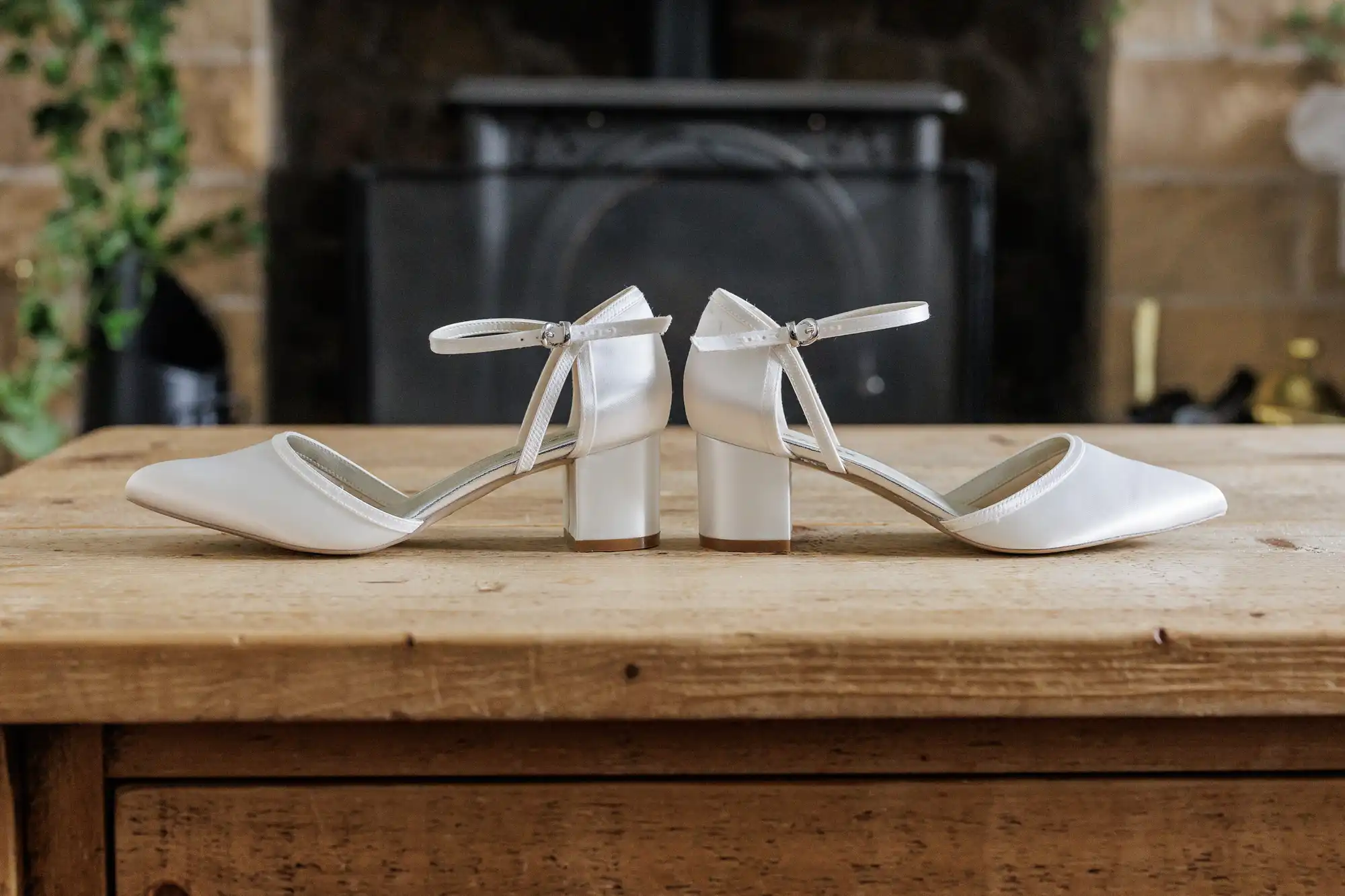A pair of white, closed-toe, block-heeled shoes with ankle straps are positioned heel-to-heel on a wooden table, in front of a blurred fireplace and stone wall background.