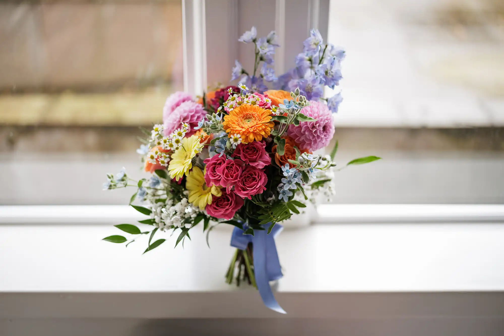 A colourful bouquet of flowers sits on a white windowsill. The bouquet includes orange, yellow, pink, and purple flowers, with greenery and small white blooms, tied together with a blue ribbon. Light streams in through the window behind it.