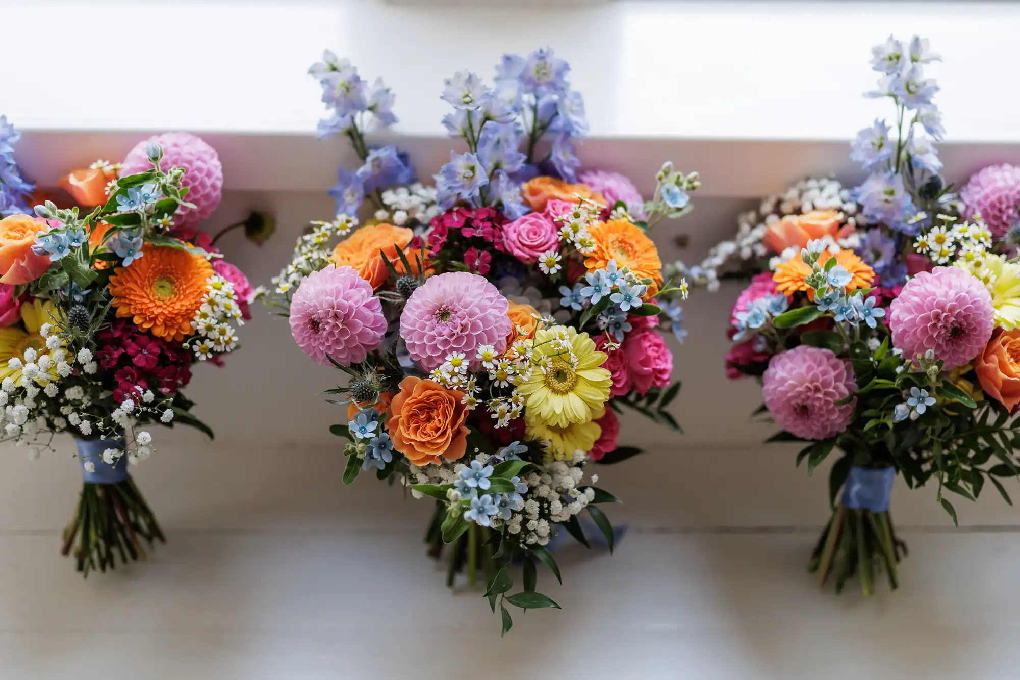 Three colourful bouquets with pink dahlias, orange roses, yellow gerbera daisies, small white and blue flowers, and greenery are arranged side by side on a white surface below a windowsill, tied with blue ribbon. Light streams in from above.