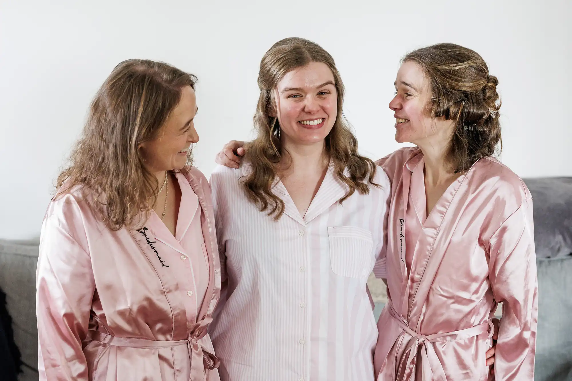Three women stand together indoors, smiling warmly. The woman in the middle wears white striped pyjamas, while the women on either side wear matching light pink satin dressing gowns and look at her affectionately. They appear relaxed and happy, likely preparing for a special occasion.