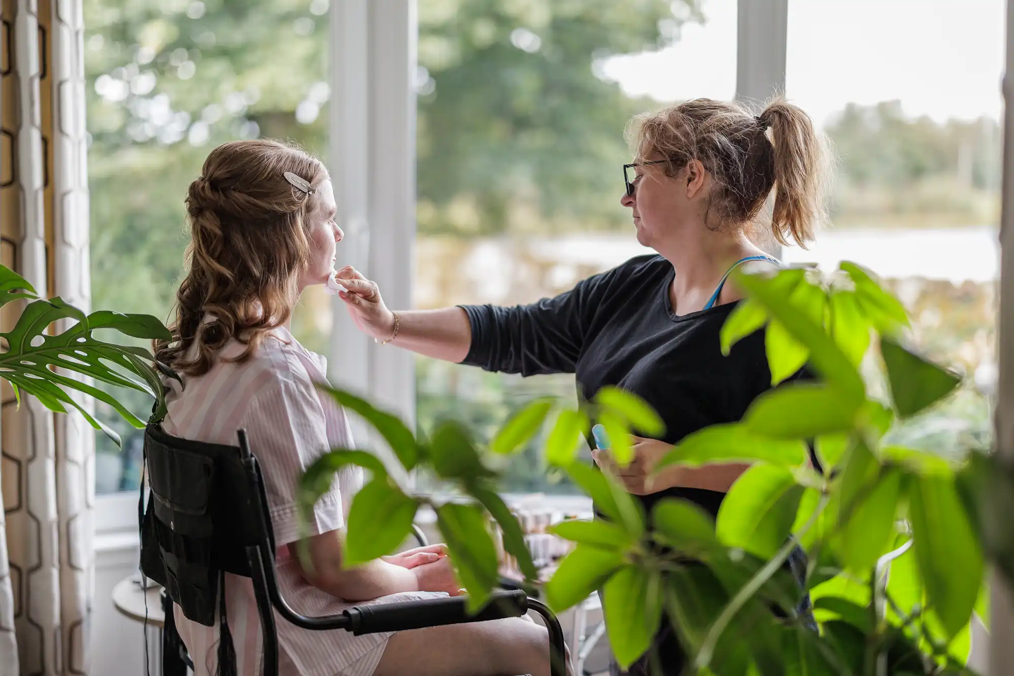 A woman applies make-up to another woman seated by a large window with greenery outside. Several leafy plants are in the foreground, partially obscuring the scene. Natural light fills the room, creating a calm, intimate atmosphere.