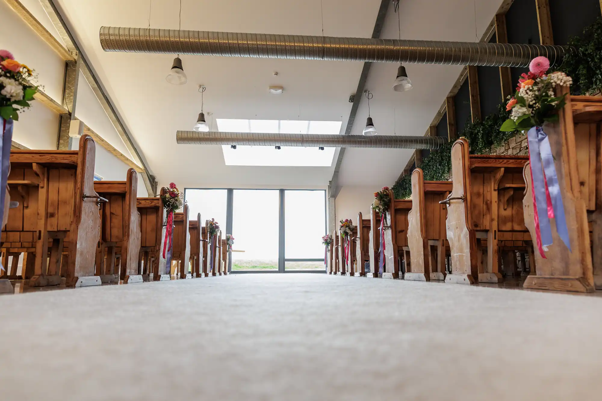 Low-angle view of a bright, modern wedding venue with wooden pews arranged in rows. Each pew is decorated with flower arrangements and colourful ribbons. Large windows at the end let natural light flood the room. Exposed ductwork is visible on the ceiling.