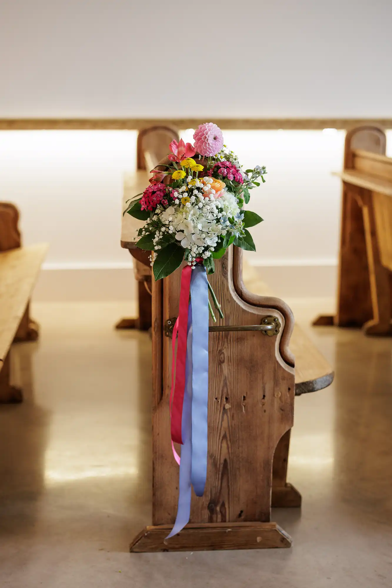 A wooden church pew decorated with a bouquet of colourful flowers, including pink, white, and yellow blooms, and green leaves. Long pink and purple ribbons hang from the bouquet. Other wooden pews are visible in the softly lit background.