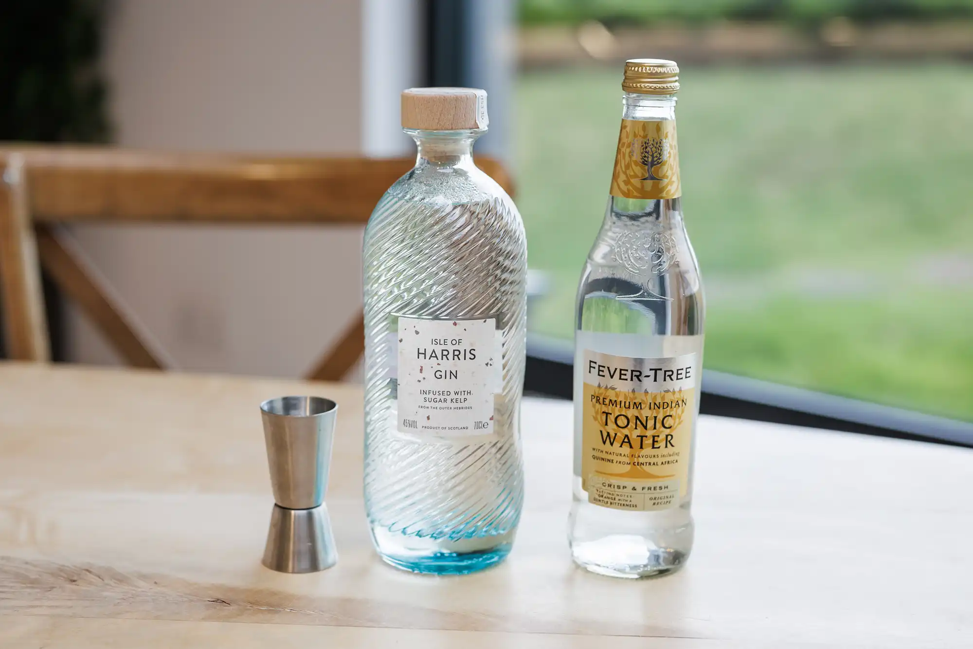 A glass bottle of Isle of Harris Gin, a bottle of Fever-Tree tonic water, and a metal measure sit on a light wooden table near a window, with a blurred green background outside.