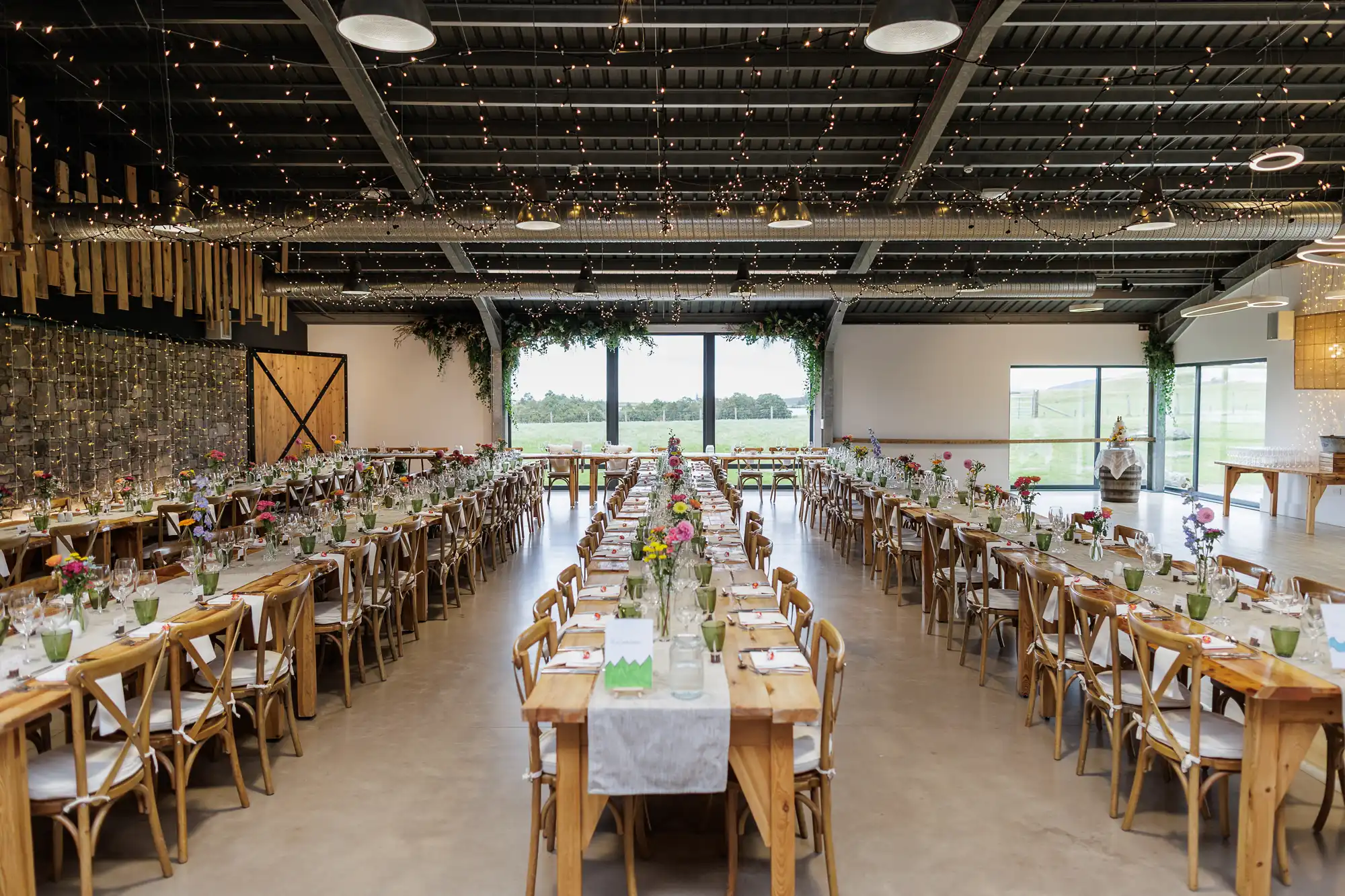 A spacious, rustic banqueting hall is set for an event with long wooden tables and chairs. Tables are adorned with white runners, flowers, and glassware. Fairy lights hang from the ceiling, and large windows at the back reveal a green landscape.