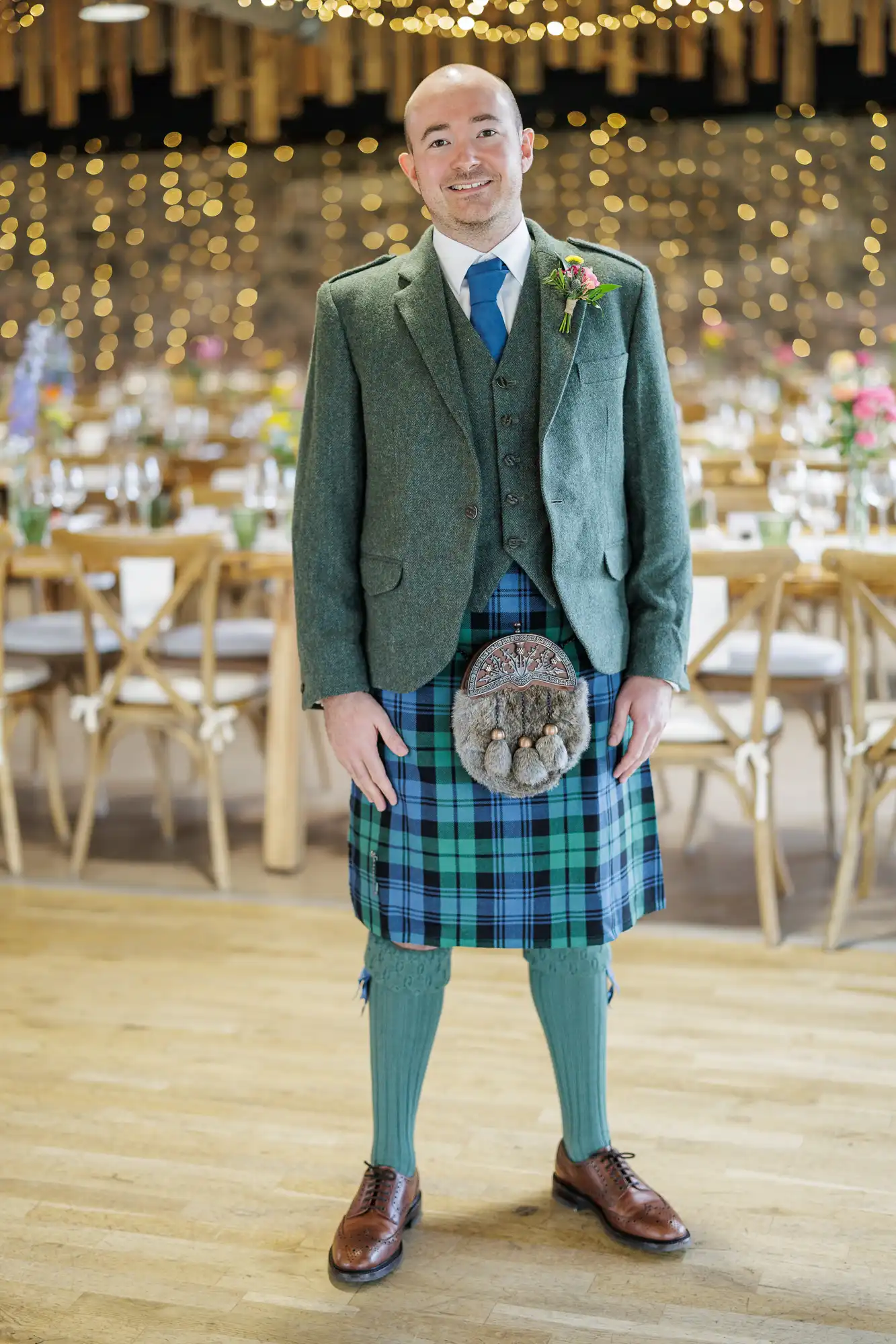 A smiling man stands indoors on a wooden floor, wearing a green tweed jacket, blue-green tartan kilt, matching socks, brown shoes, blue tie, and buttonhole. Behind him are decorated tables and warm fairy lights.