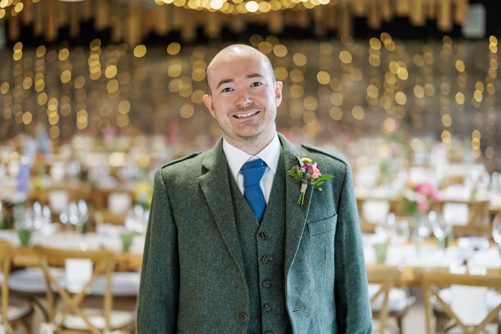 A smiling man in a green tweed suit and blue tie stands indoors at a formal event. He has a buttonhole on his lapel. Behind him are round tables with white tablecloths, wooden chairs, colourful flowers, and warm fairy lights in the background.