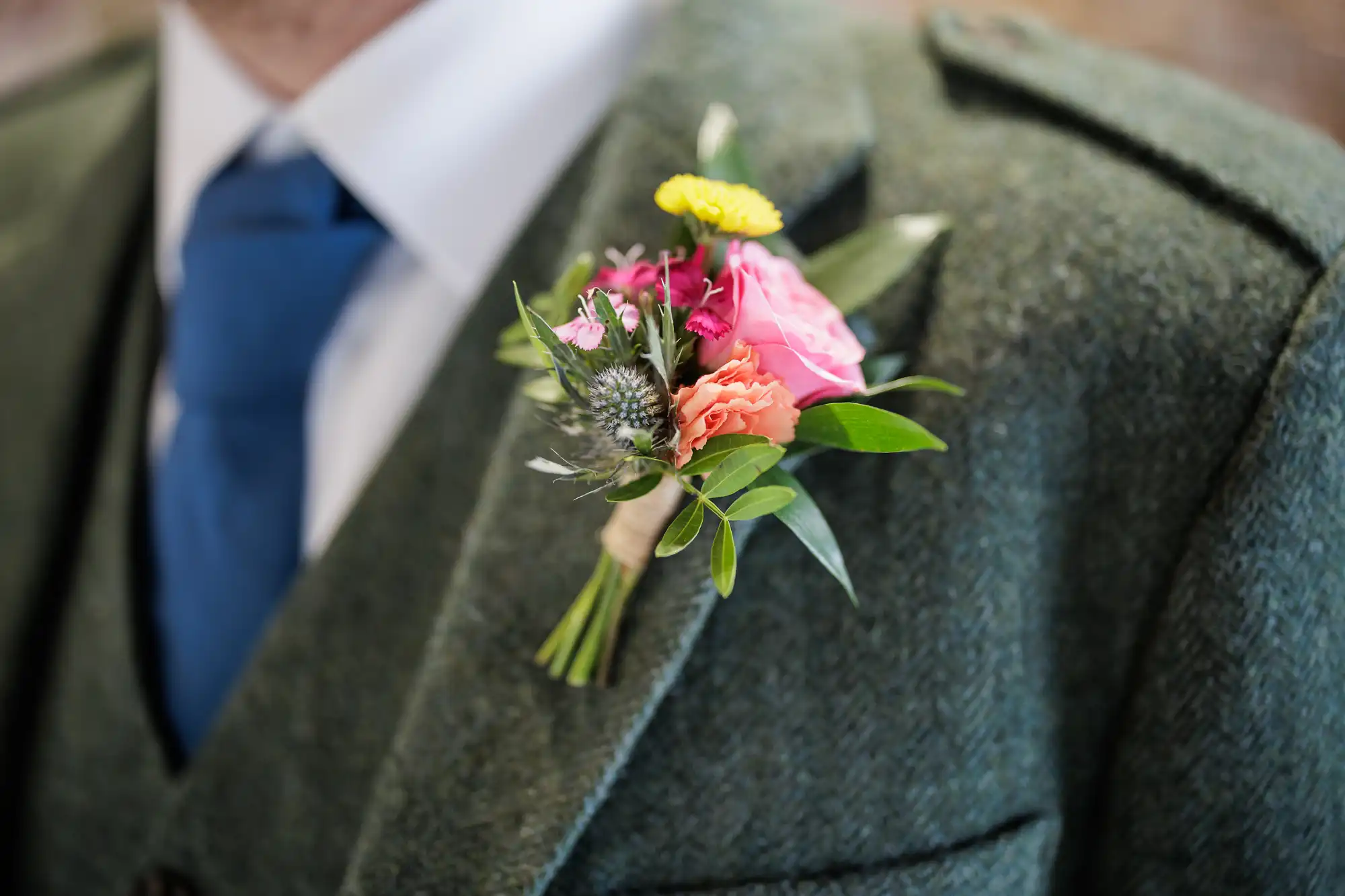 A close-up of a person wearing a green tweed suit, white shirt, and blue tie. A colourful buttonhole with pink, yellow, orange, and purple flowers and green leaves is pinned to the lapel. The face is not visible.