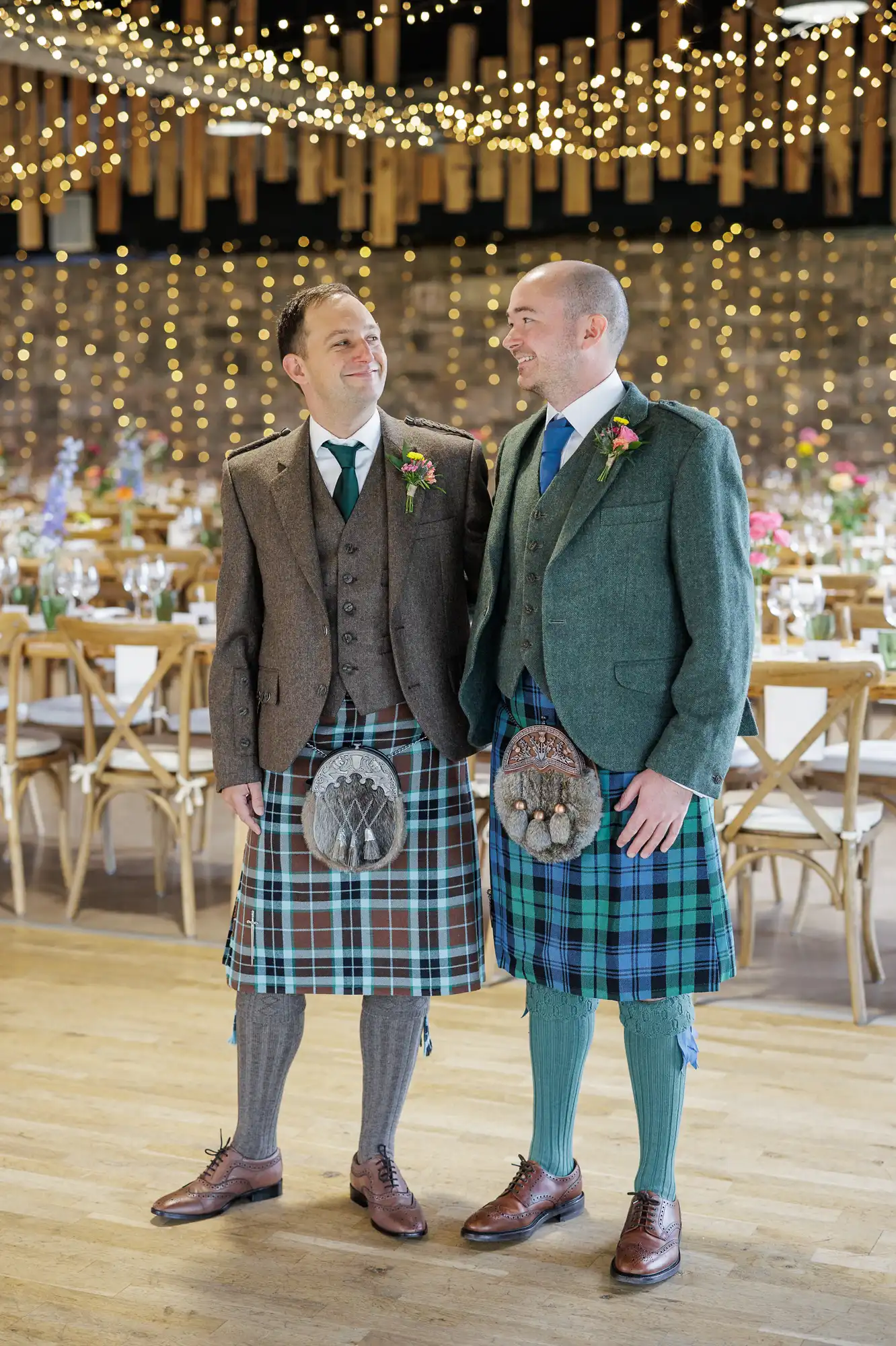 Two men in kilts, jackets, and ties stand smiling at each other in a decorated banquet hall with wooden tables and twinkling fairy lights above. Both wear sporrans; one has blue and green tartan, the other brown and blue. The mood is festive and joyful.