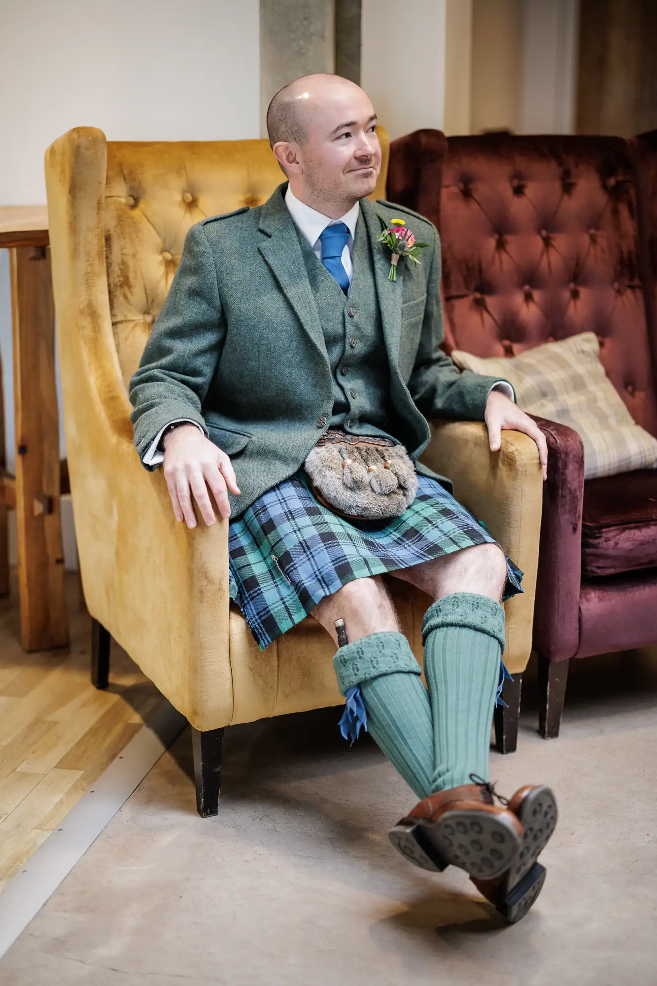 A man wearing a green jacket, blue tartan kilt, green socks, and brown shoes sits on a yellow armchair. He has a bald head, is smiling slightly, and wears a buttonhole and sporran. A burgundy chair and checked cushion are in the background.