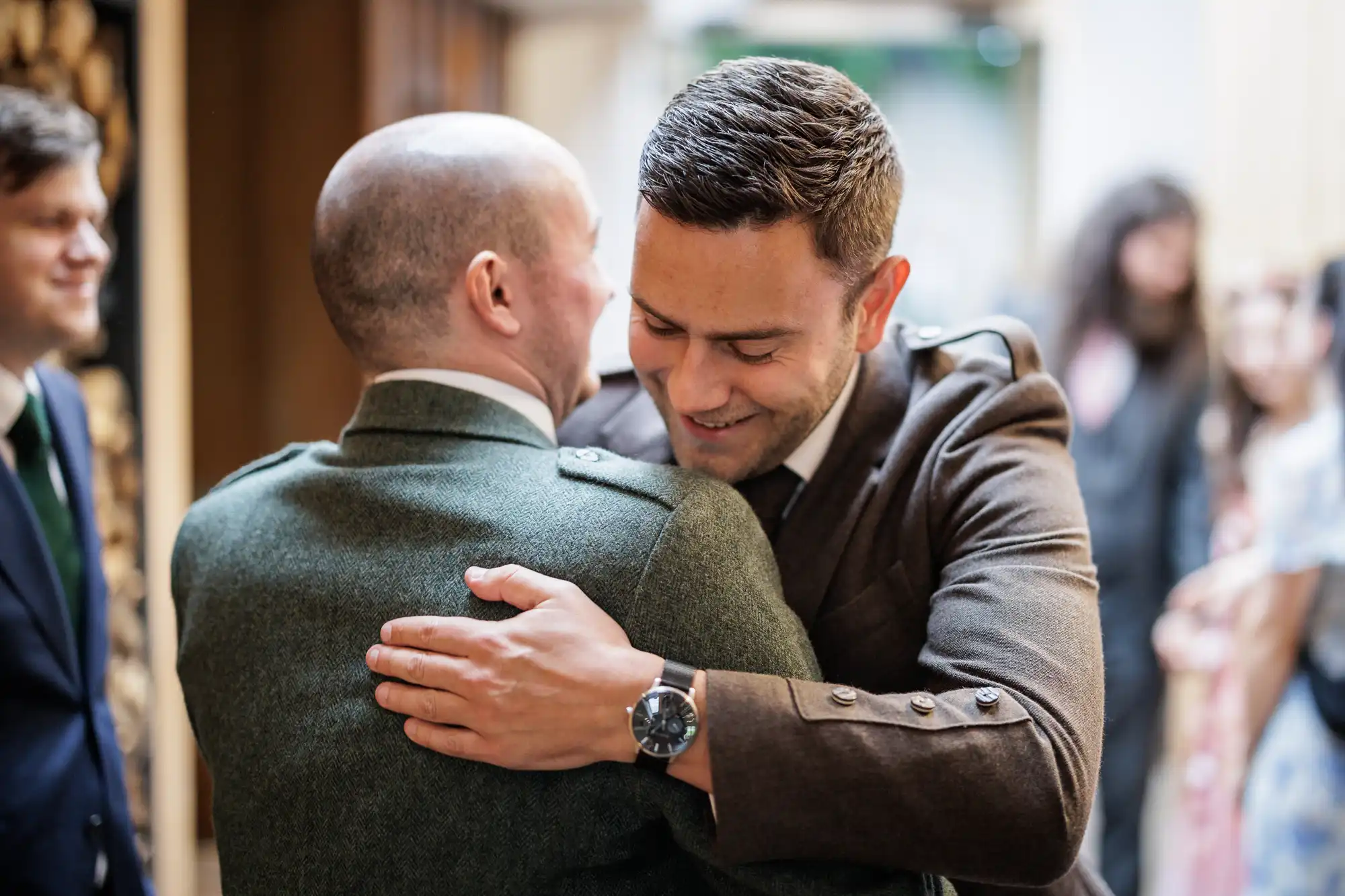 Two men in formal attire warmly embrace each other, both smiling. The man facing the camera has short dark hair and wears a brown suit, whilst the other has a shaven head and wears a green jacket. Blurred people are visible in the background.