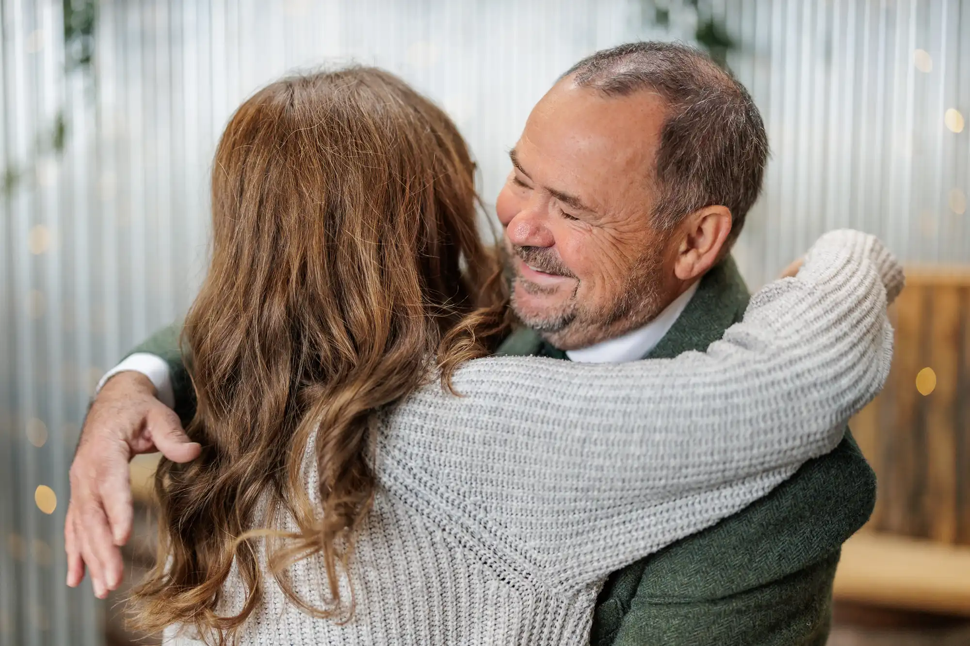 A smiling older man with short grey hair and a trimmed beard warmly hugs a woman with long brown hair, both wearing cosy jumpers, in a bright, softly lit room with blurred, festive lights in the background.