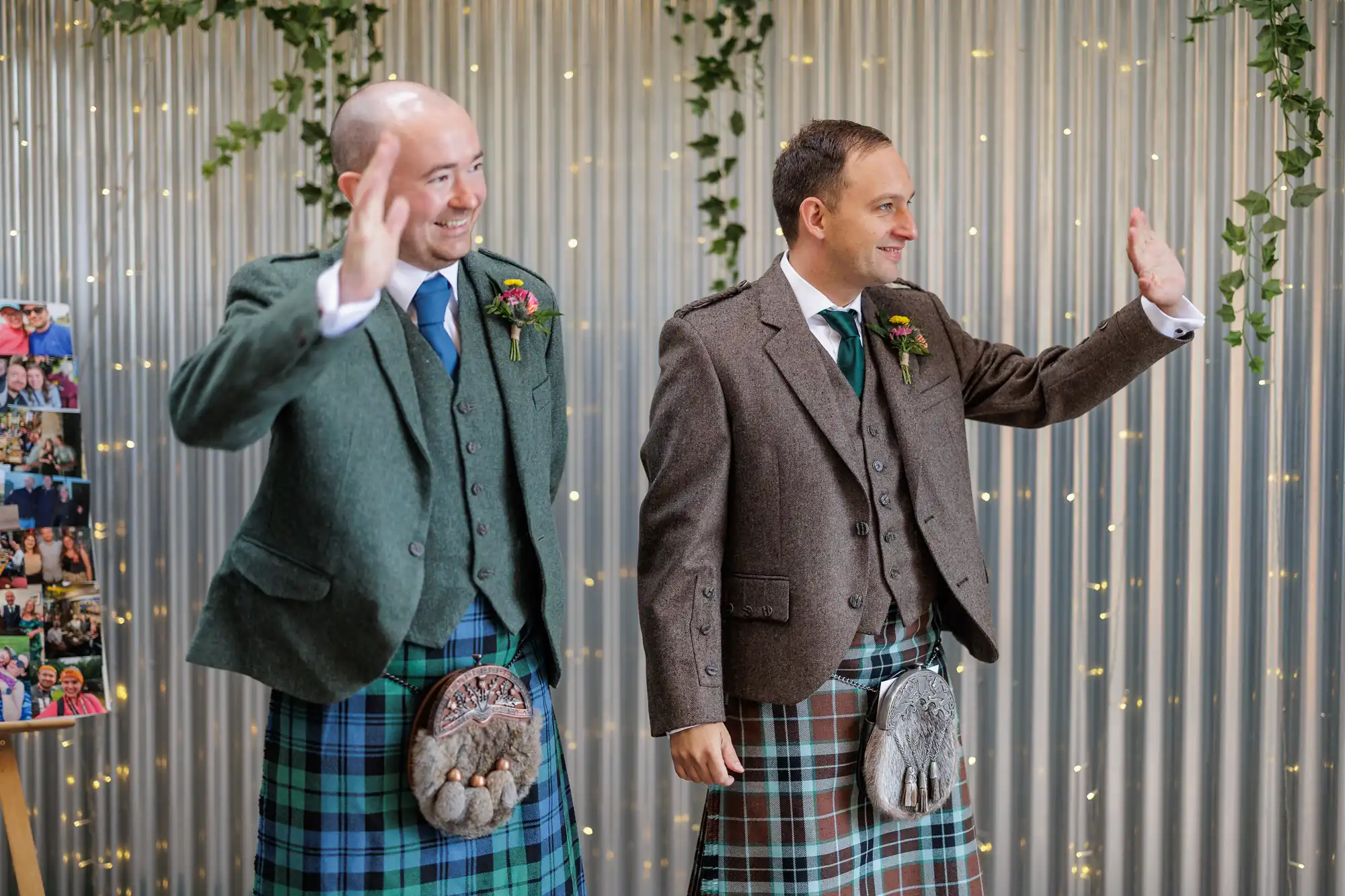 Two men in jackets, ties, and tartan kilts with sporrans stand side by side, smiling and waving. Behind them is a silver corrugated wall decorated with fairy lights and hanging greenery. At left, part of a photo collage is visible.
