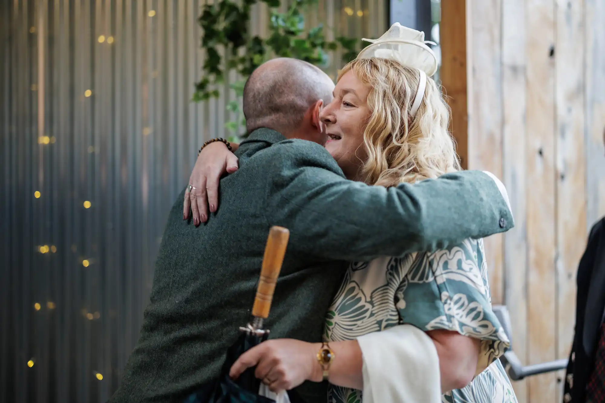A man and woman hug warmly indoors. The man, seen from behind, holds an umbrella. The woman, with curly blonde hair and a white hat, smiles. She wears a blue patterned dress and cream shawl. Fairy lights and greenery decorate the metal wall in the background.