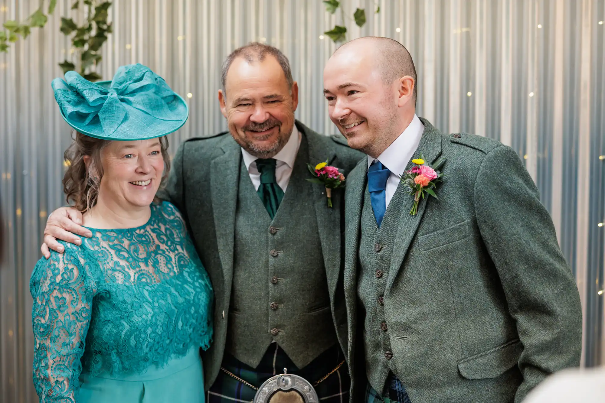 Three adults smile together at an event. Two men wear green tweed jackets with buttonholes and tartan kilts; the woman wears a teal lace dress and a matching hat. They stand in front of a corrugated metal wall decorated with greenery and fairy lights.