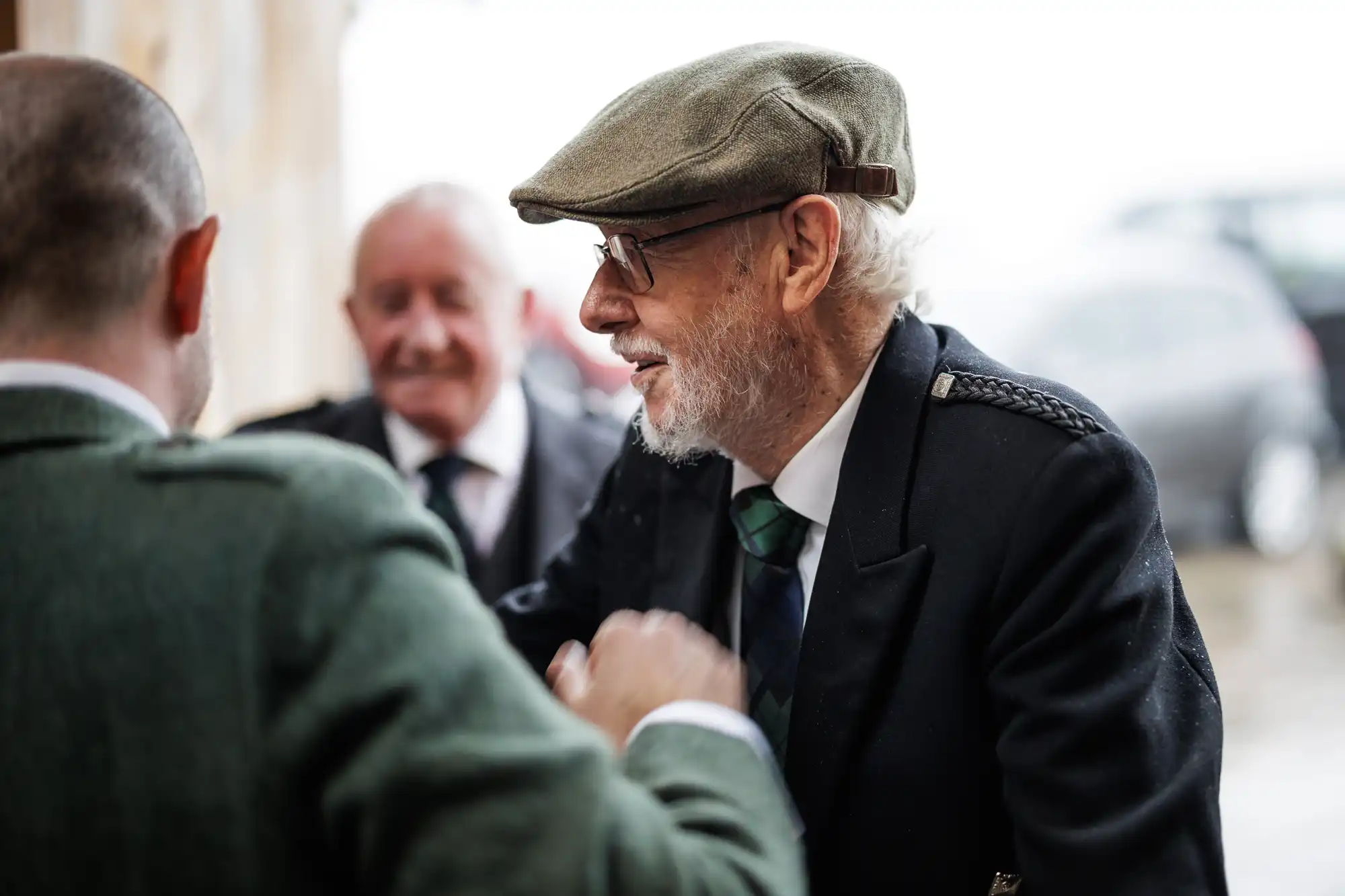 An older man with a grey beard and flat cap, wearing glasses, a dark overcoat, and a green checked tie, is smiling and shaking hands with another man in a green jacket. A third man in a suit smiles in the blurred background.