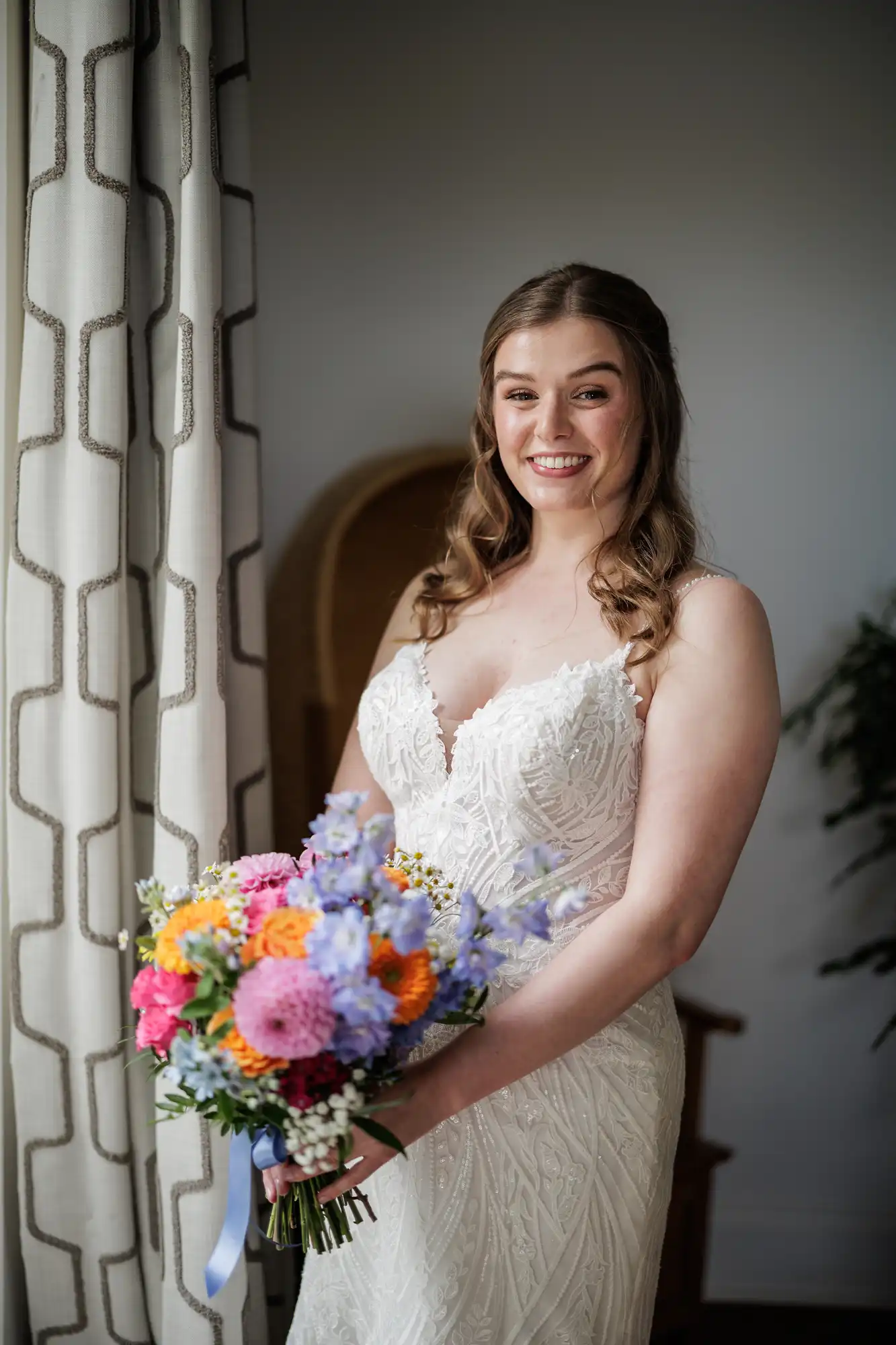 A smiling bride with wavy brown hair in a white lace wedding dress holds a colourful bouquet of pink, orange, purple, and blue flowers. She stands indoors by a window with patterned curtains, with soft natural light illuminating her face.