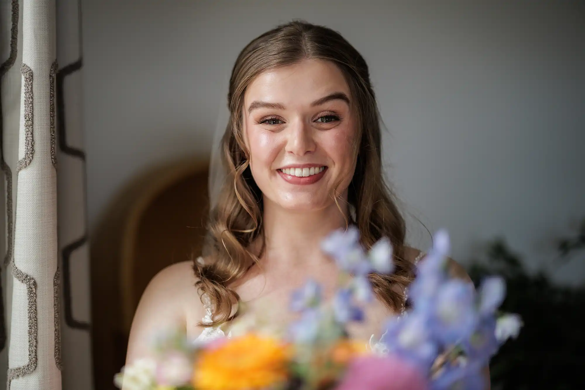 A young woman with long, wavy light brown hair smiles brightly, standing indoors near a window with patterned curtains. She holds a colourful bouquet of flowers in the foreground, slightly out of focus. She wears a white dress and appears joyful.